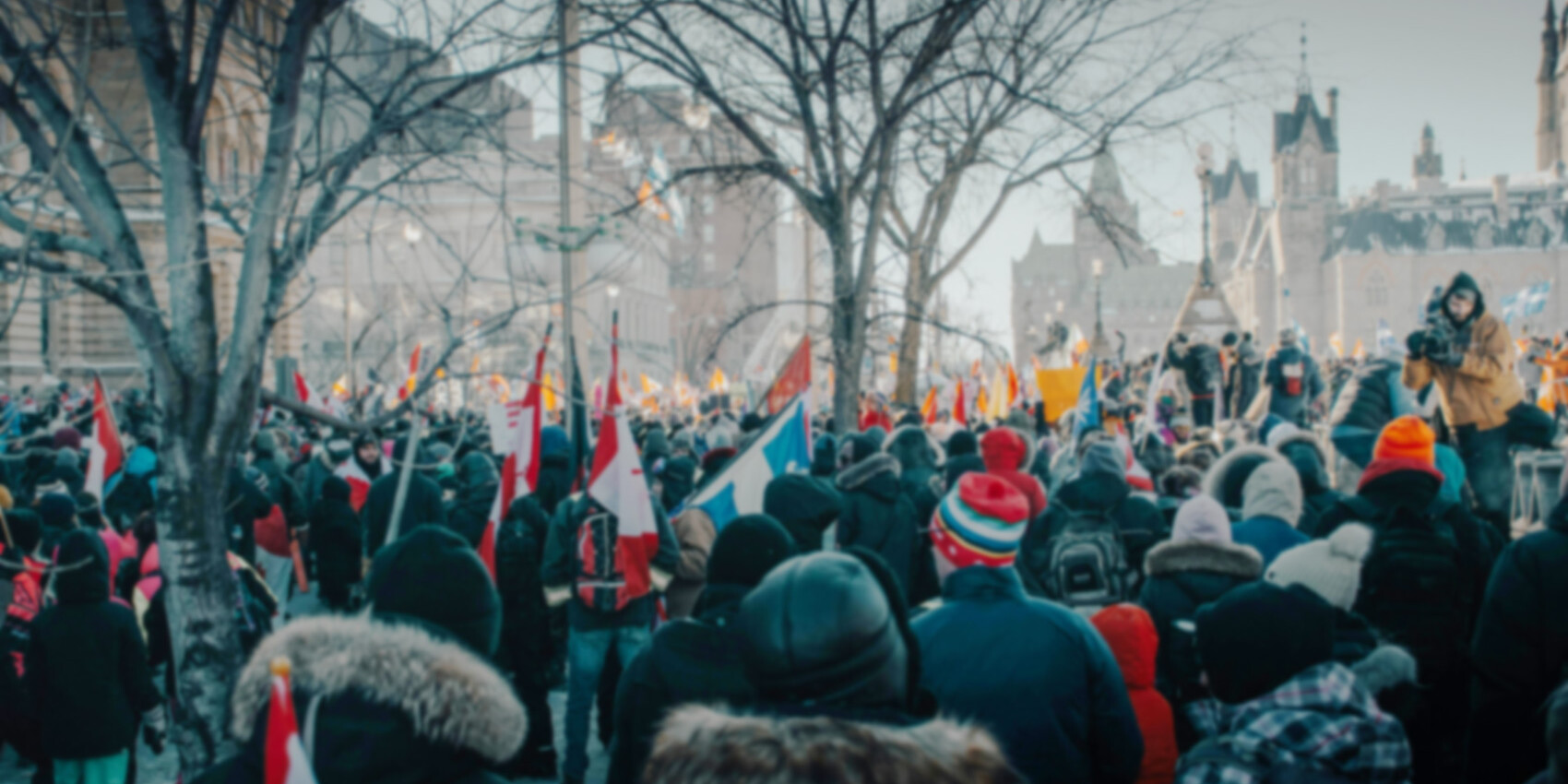 A large crowd gathers outdoors in winter, many wearing coats and hats. People hold Canadian flags and banners, demonstrating their influence in Canadian politics. Leafless trees and historic buildings are visible in the background.