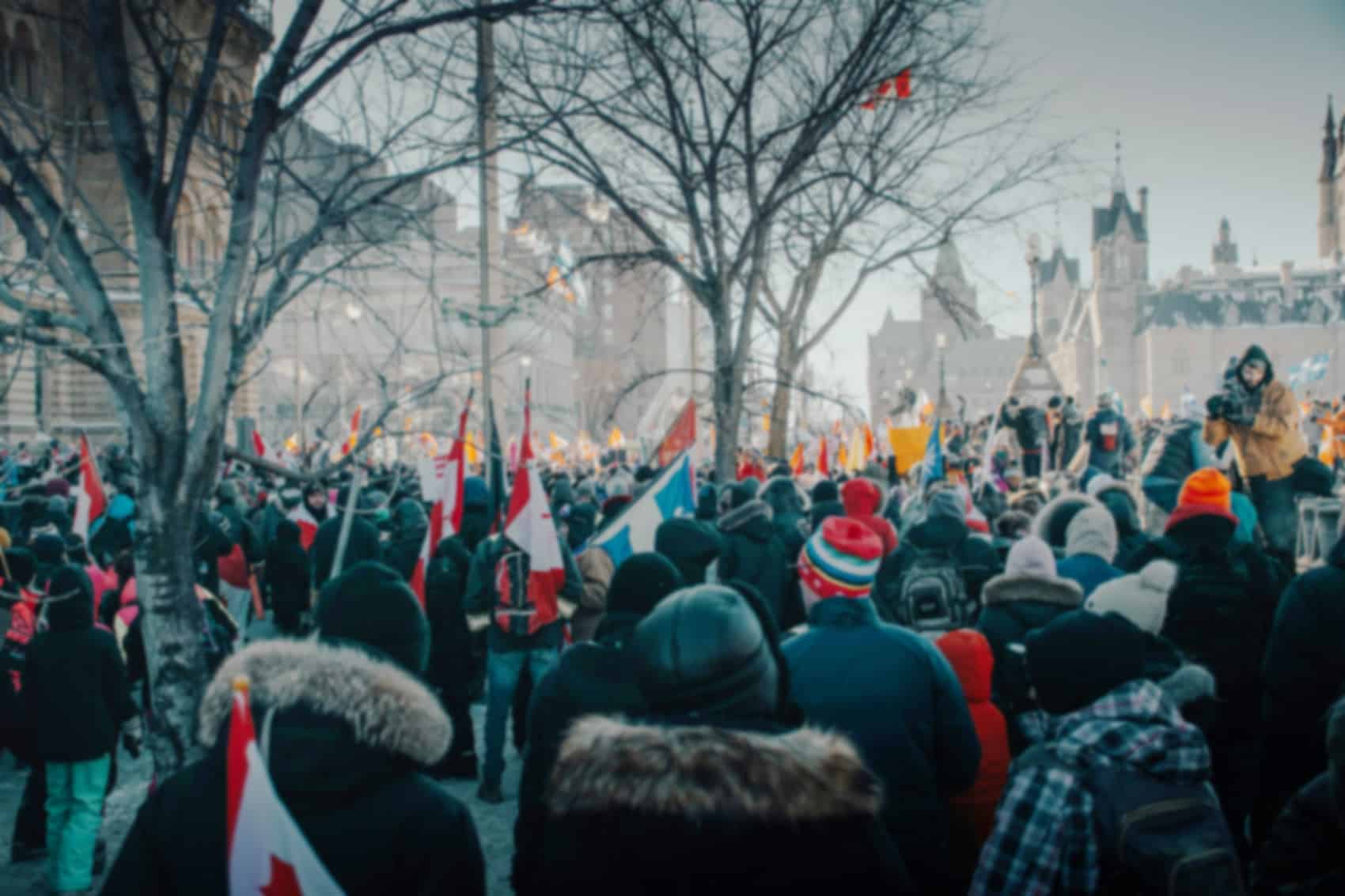 A large crowd gathers outdoors in winter, many wearing coats and hats. People hold Canadian flags and banners, demonstrating their influence in Canadian politics. Leafless trees and historic buildings are visible in the background.