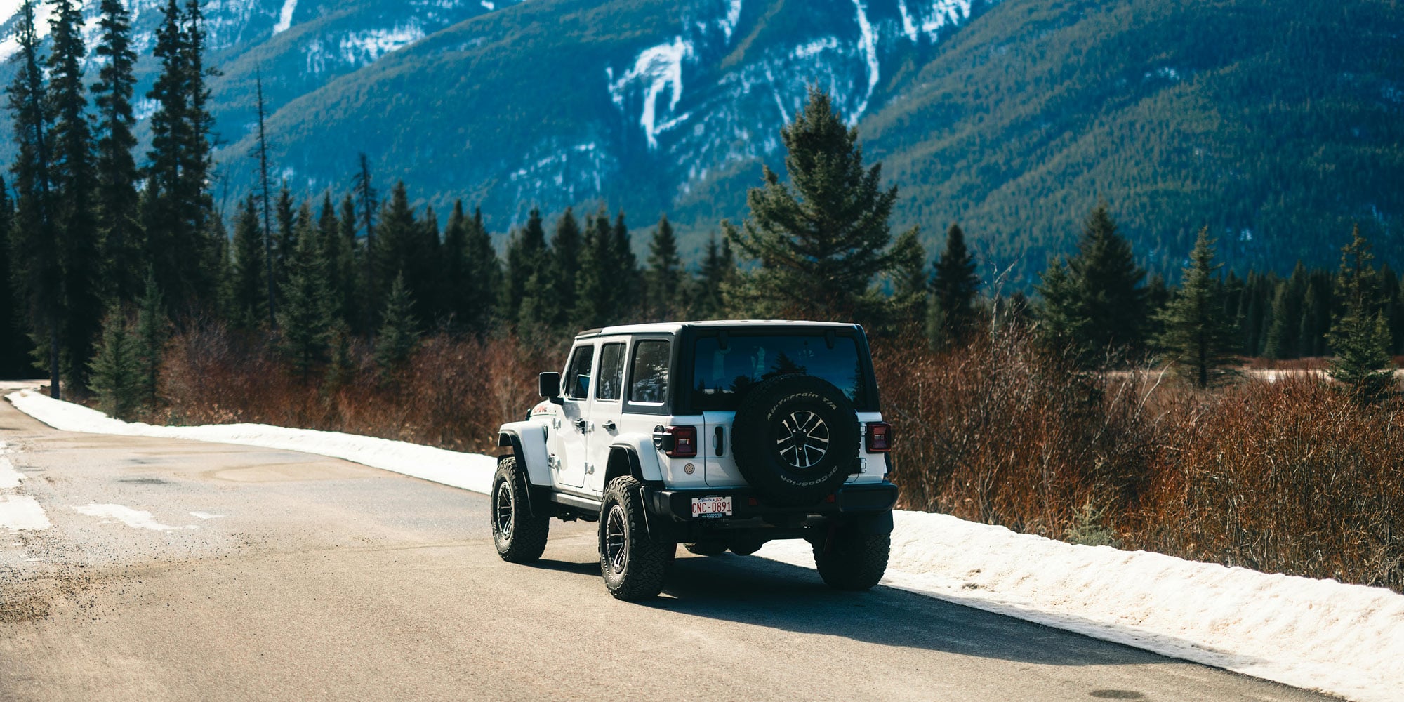 A white SUV is parked on a snow-lined mountain road surrounded by evergreen trees, with tall, snowy mountains in the background—reflecting landscapes where a majority of Canadians support equalization, even in Alberta, under the clear sky.