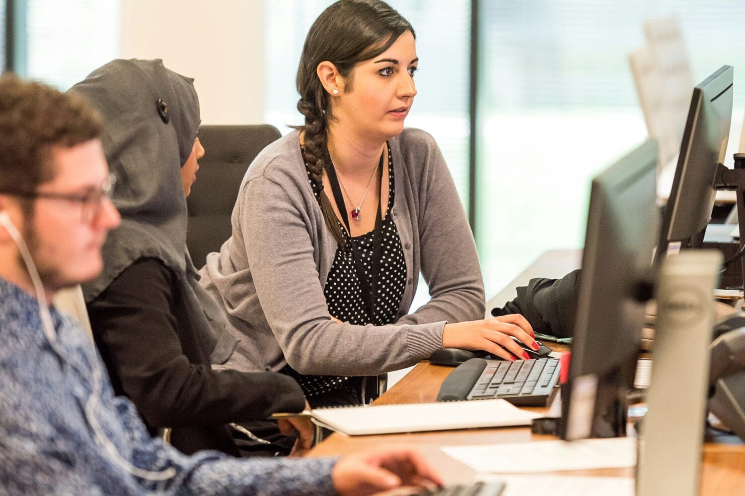 Three people work at desks in an office—one woman types, while another woman in a headscarf and a man with glasses and earphones focus on their screens, reflecting how greater inclusion is a win-win strategy for the recovery.