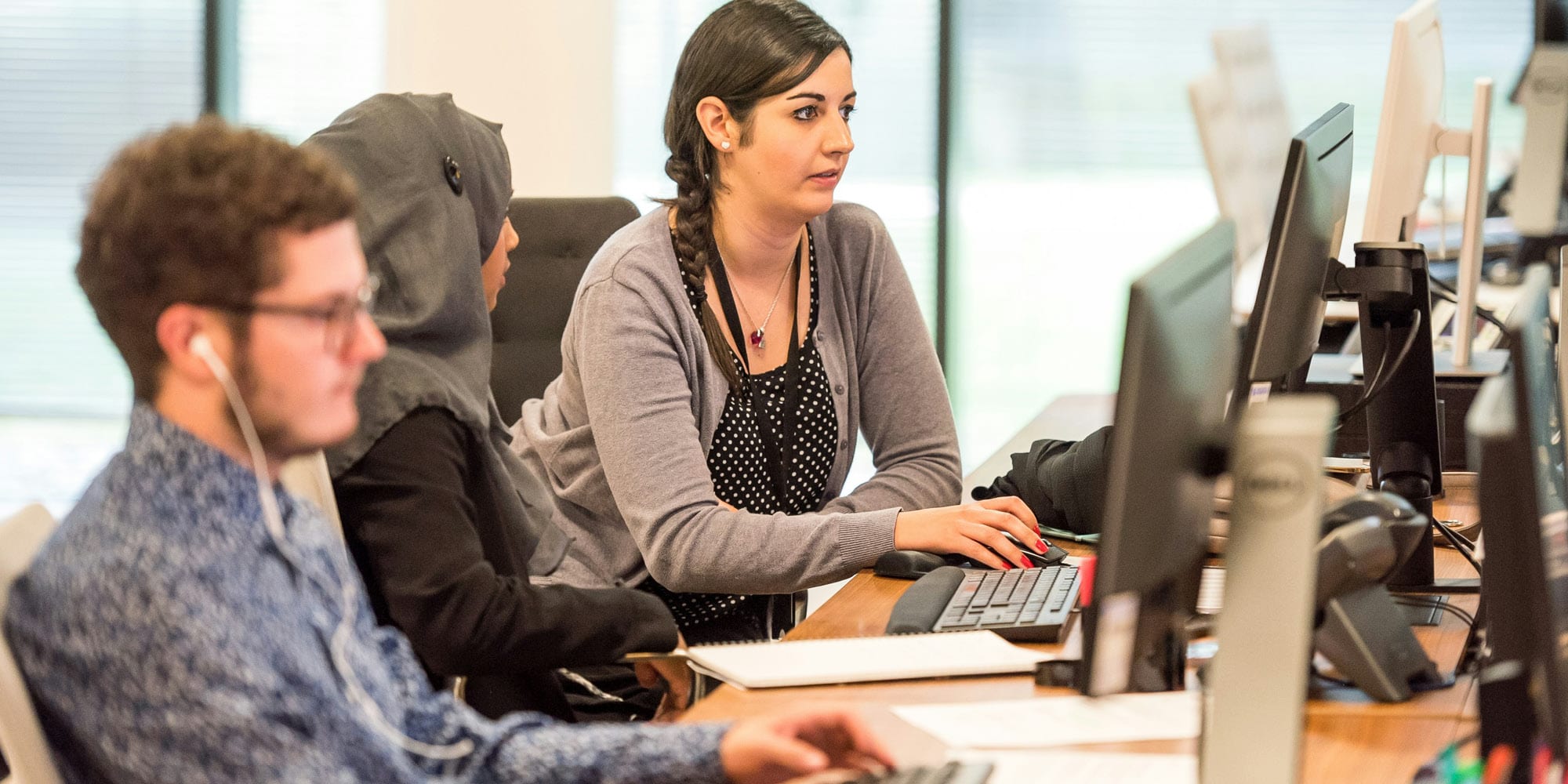 Three people work at desks in an office—one woman types, while another woman in a headscarf and a man with glasses and earphones focus on their screens, reflecting how greater inclusion is a win-win strategy for the recovery.