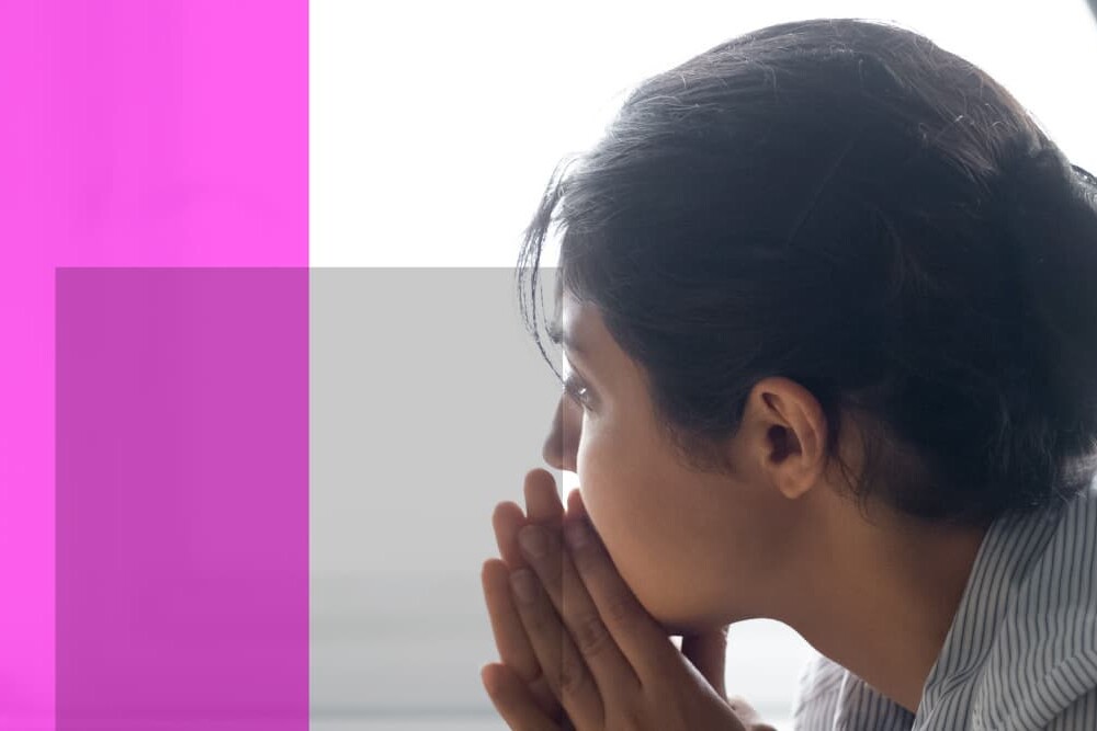 A person with dark hair tied back rests their chin on clasped hands, looking thoughtful or concerned—perhaps reflecting on the Mind and Body: Impact of the Pandemic on Physical and Mental Health—while sitting indoors near bright light. Pink and purple translucent rectangles overlay the left.