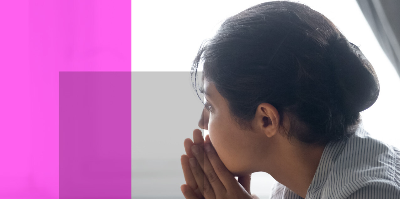 A person with dark hair tied back rests their chin on clasped hands, looking thoughtful or concerned—perhaps reflecting on the Mind and Body: Impact of the Pandemic on Physical and Mental Health—while sitting indoors near bright light. Pink and purple translucent rectangles overlay the left.