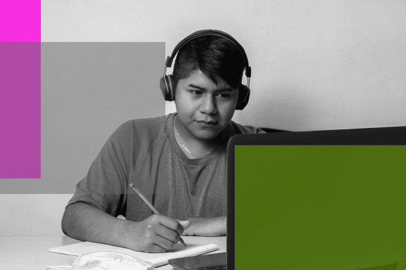 A young person wearing headphones sits at a desk, looking at a laptop screen and taking notes with a pencil—an image reflecting 