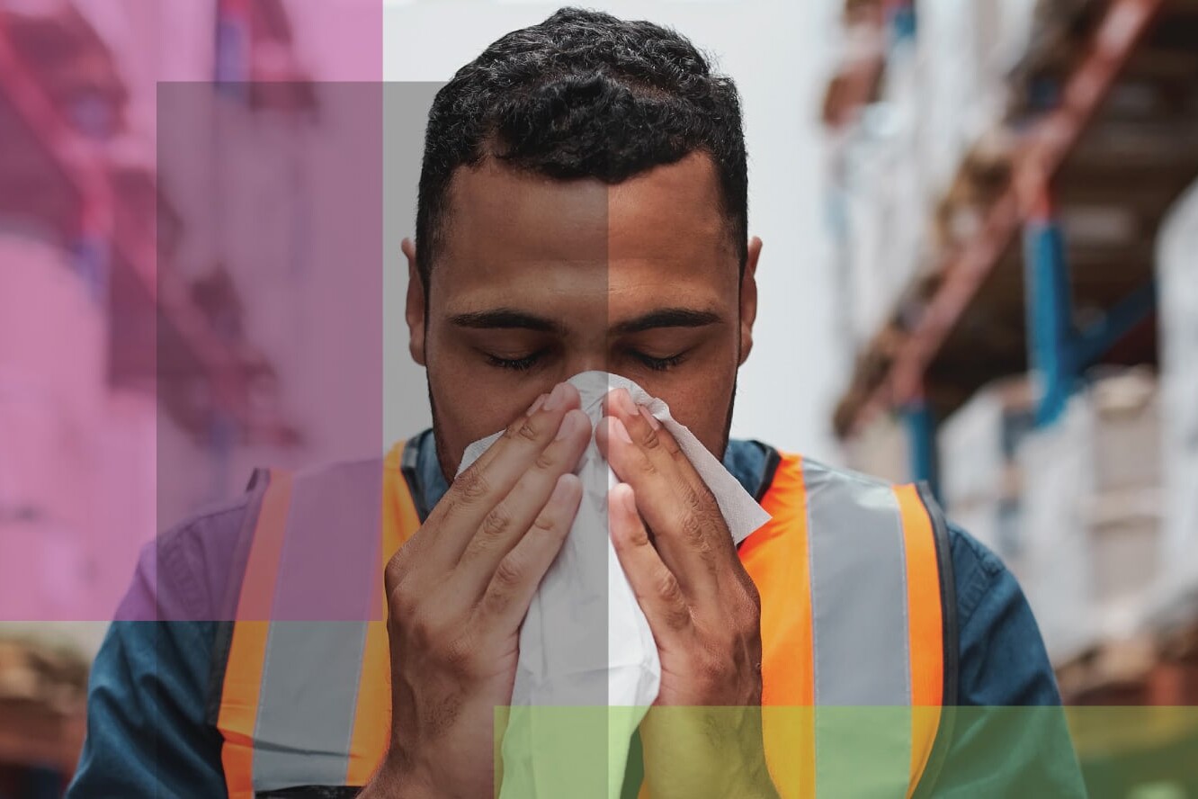 A man wearing a safety vest stands in a warehouse, holding a tissue to his nose as if sneezing—highlighting issues around working when sick and how workplace regulations will impact the post-pandemic recovery. Semi-transparent colored rectangles overlay the image.