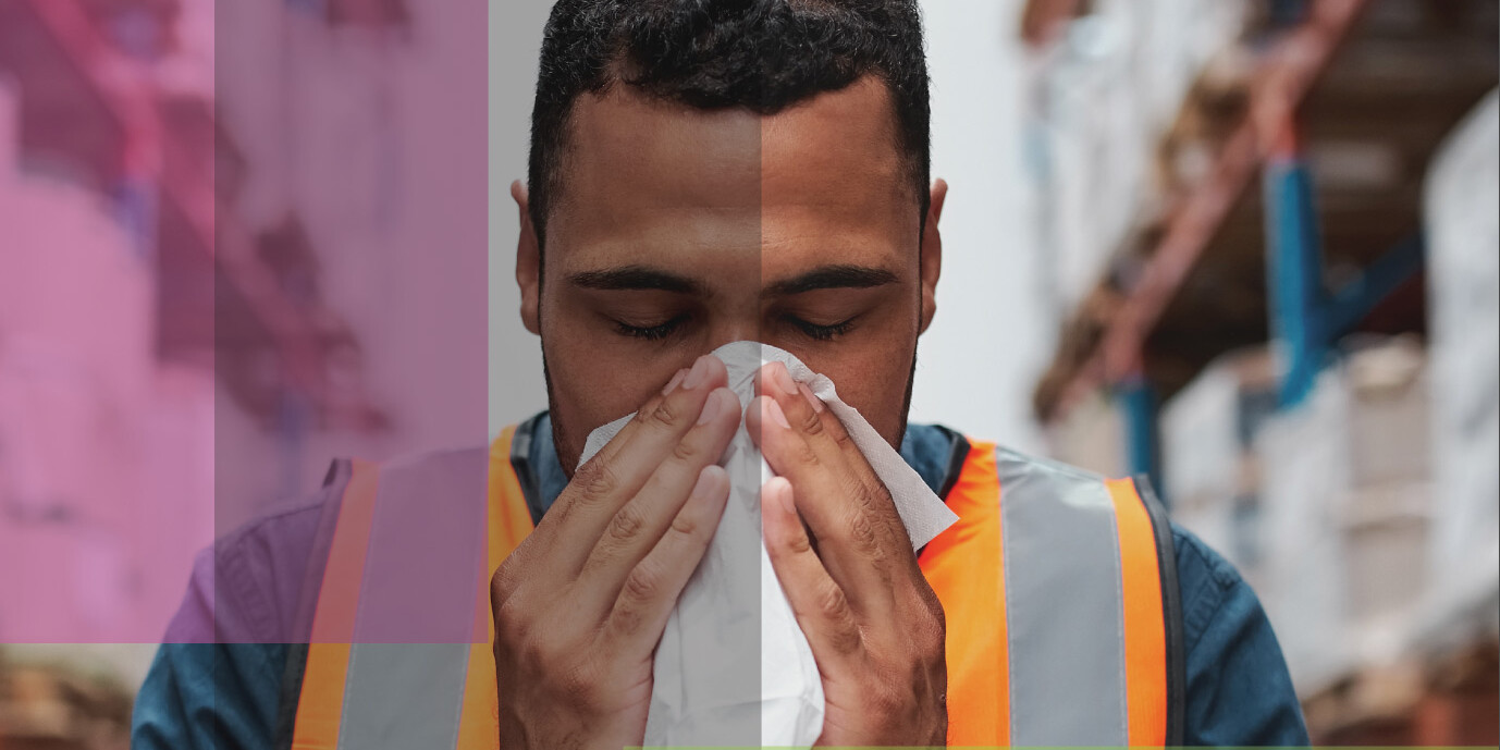 A man wearing a safety vest stands in a warehouse, holding a tissue to his nose as if sneezing—highlighting issues around working when sick and how workplace regulations will impact the post-pandemic recovery. Semi-transparent colored rectangles overlay the image.