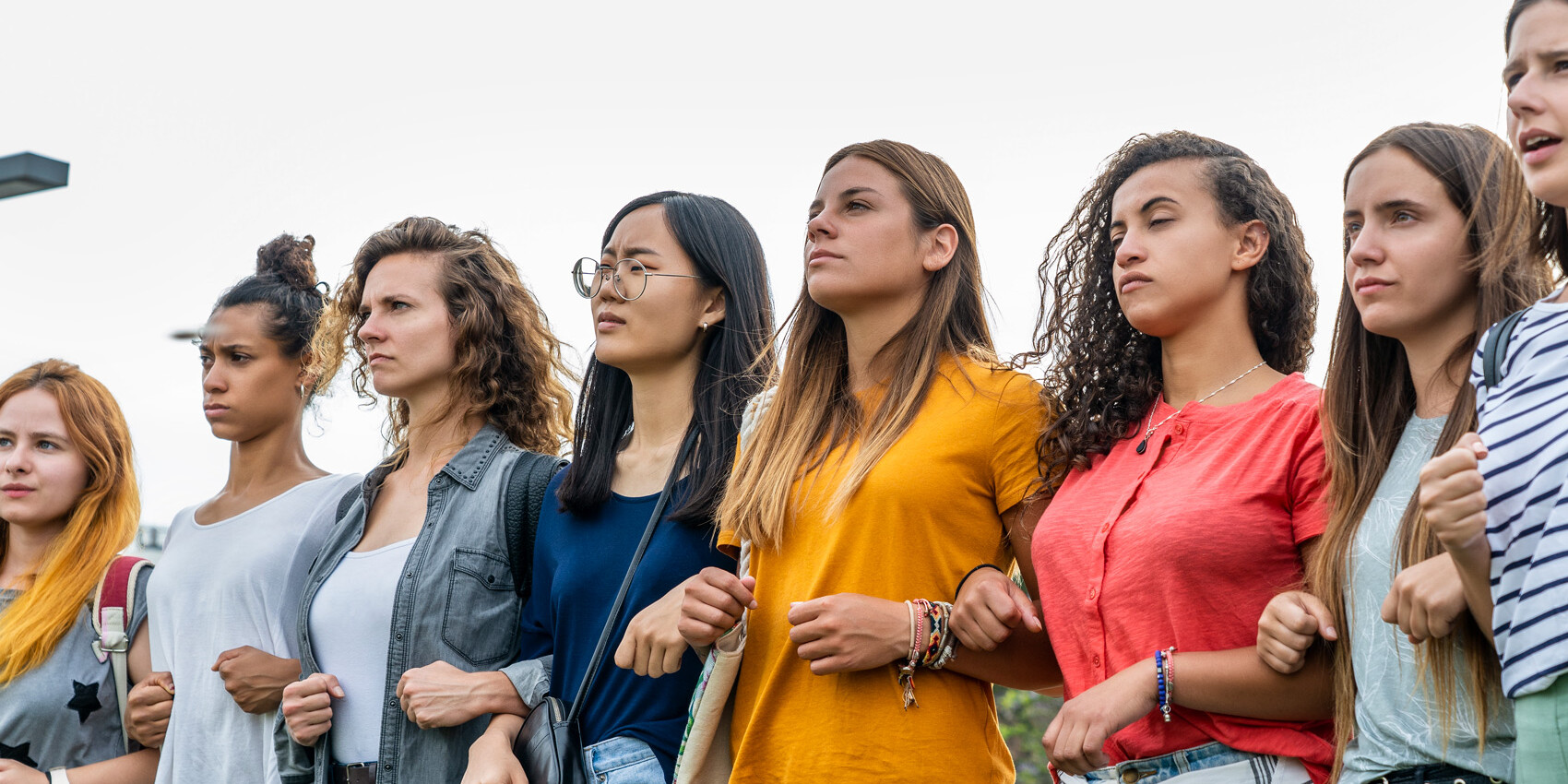 A diverse group of young women standing in a line outdoors, linking arms and looking determinedly ahead, united in solidarity for Women's Equality and the Women's Movement: an Update.