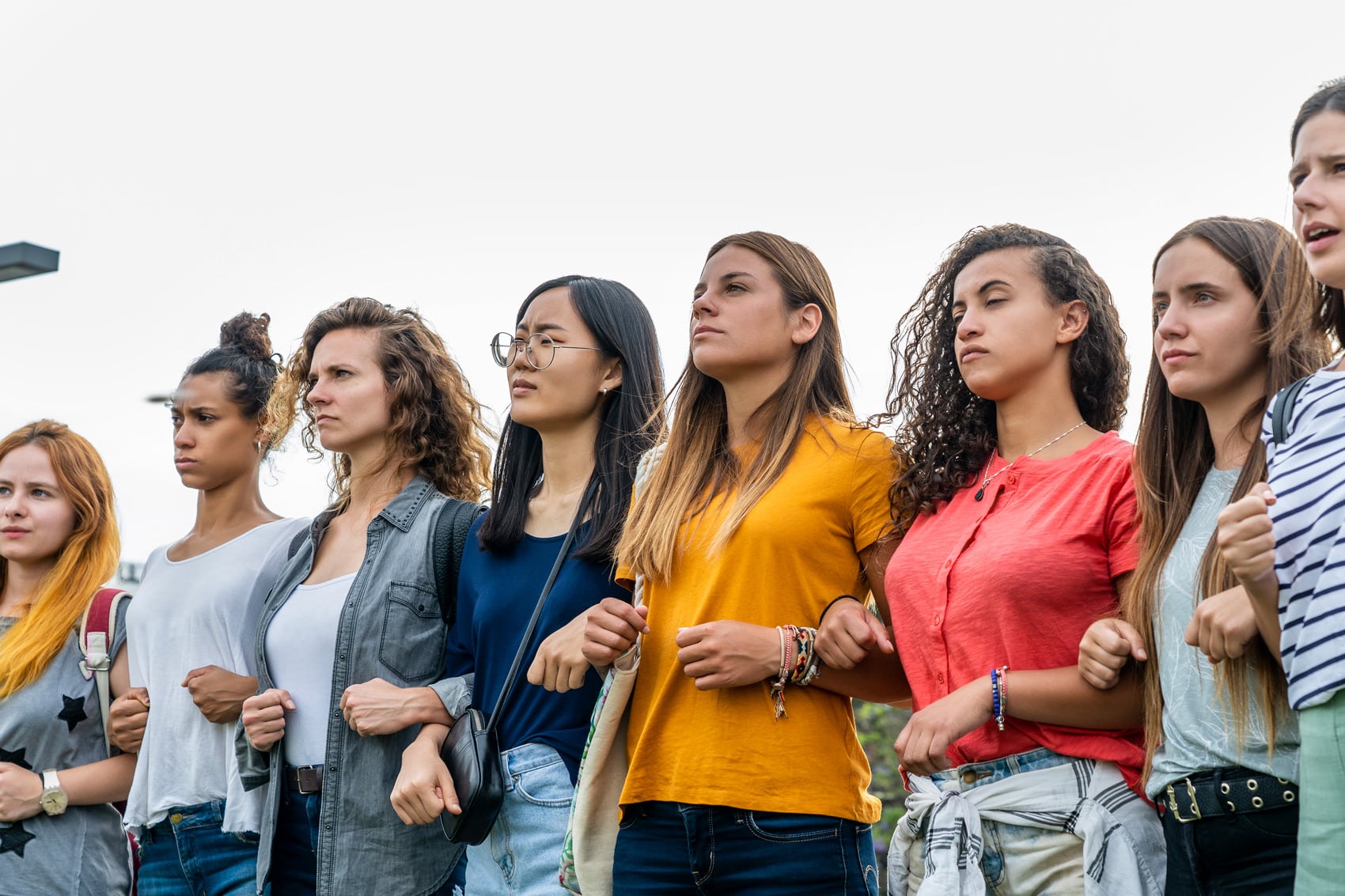 A diverse group of young women standing in a line outdoors, linking arms and looking determinedly ahead, united in solidarity for Women's Equality and the Women's Movement: an Update.