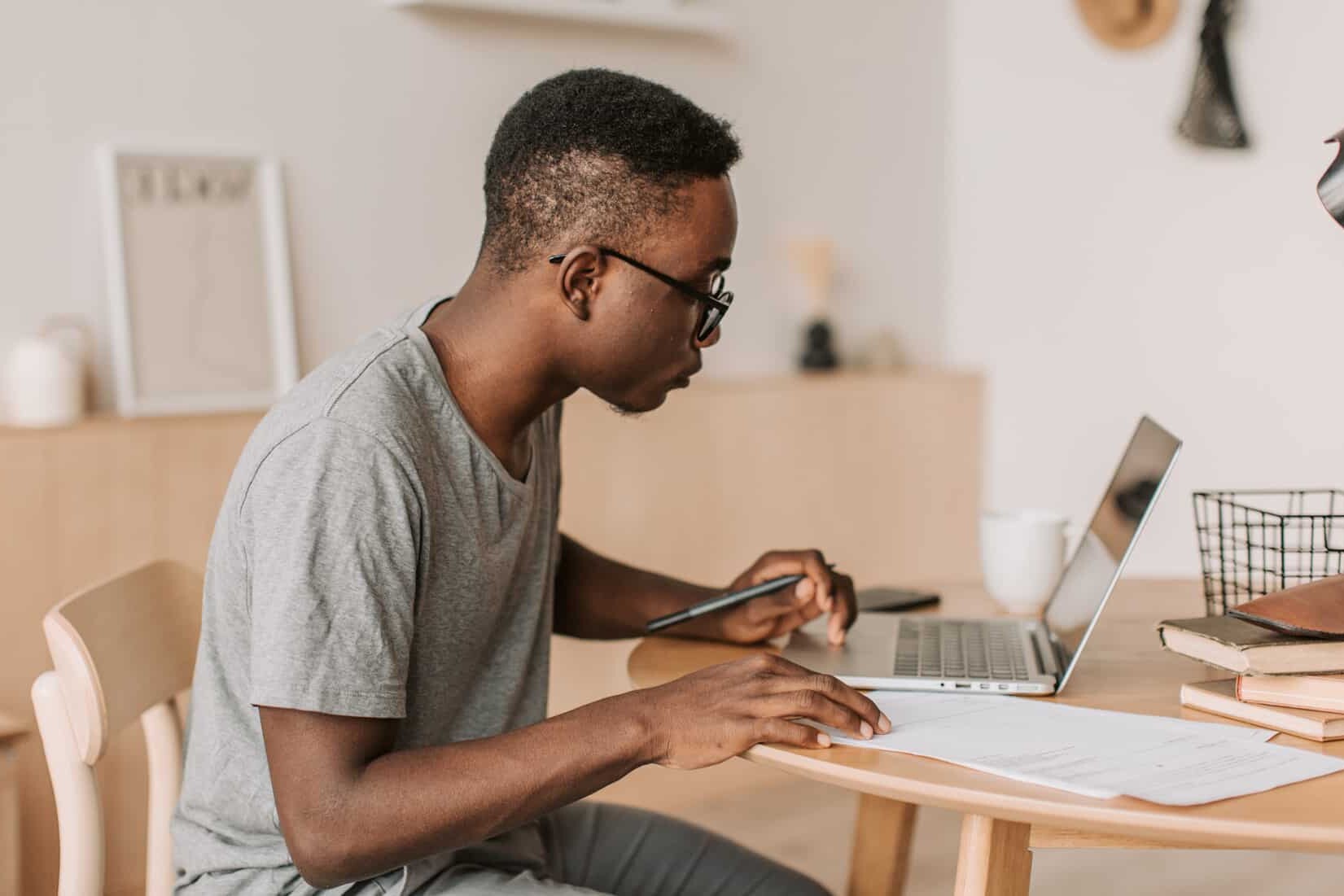 A young man wearing glasses and a gray t-shirt sits at a table, working on a laptop with papers and a pen in hand, reflecting on lessons learned: the pandemic and learning from home in Canada, concentrating in a bright, modern room.