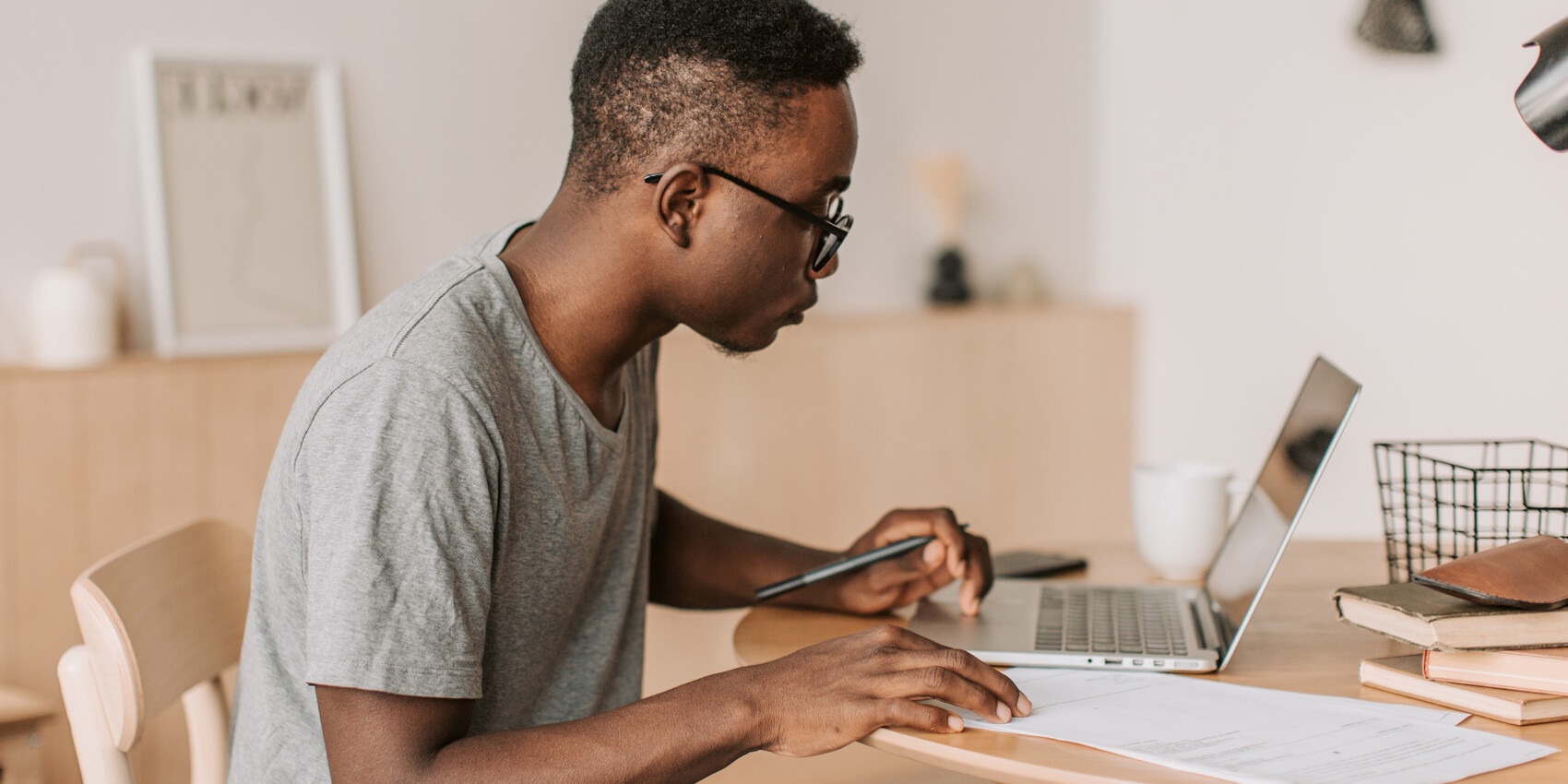 A young man wearing glasses and a gray t-shirt sits at a table, working on a laptop with papers and a pen in hand, reflecting on lessons learned: the pandemic and learning from home in Canada, concentrating in a bright, modern room.