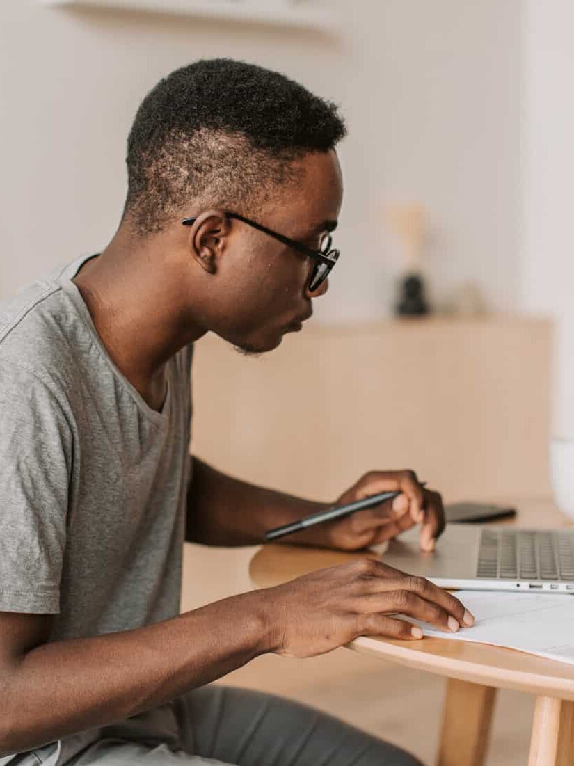 A young man wearing glasses and a gray t-shirt sits at a table, working on a laptop with papers and a pen in hand, reflecting on lessons learned: the pandemic and learning from home in Canada, concentrating in a bright, modern room.