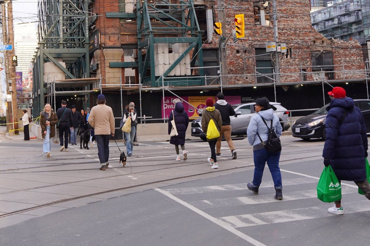 A group of people cross a city street at a crosswalk near a construction site, some carrying green shopping bags. Buildings and traffic lights are visible, reflecting how Canadians aren’t just adapting to diversity – there are data to show we’re embracing it.