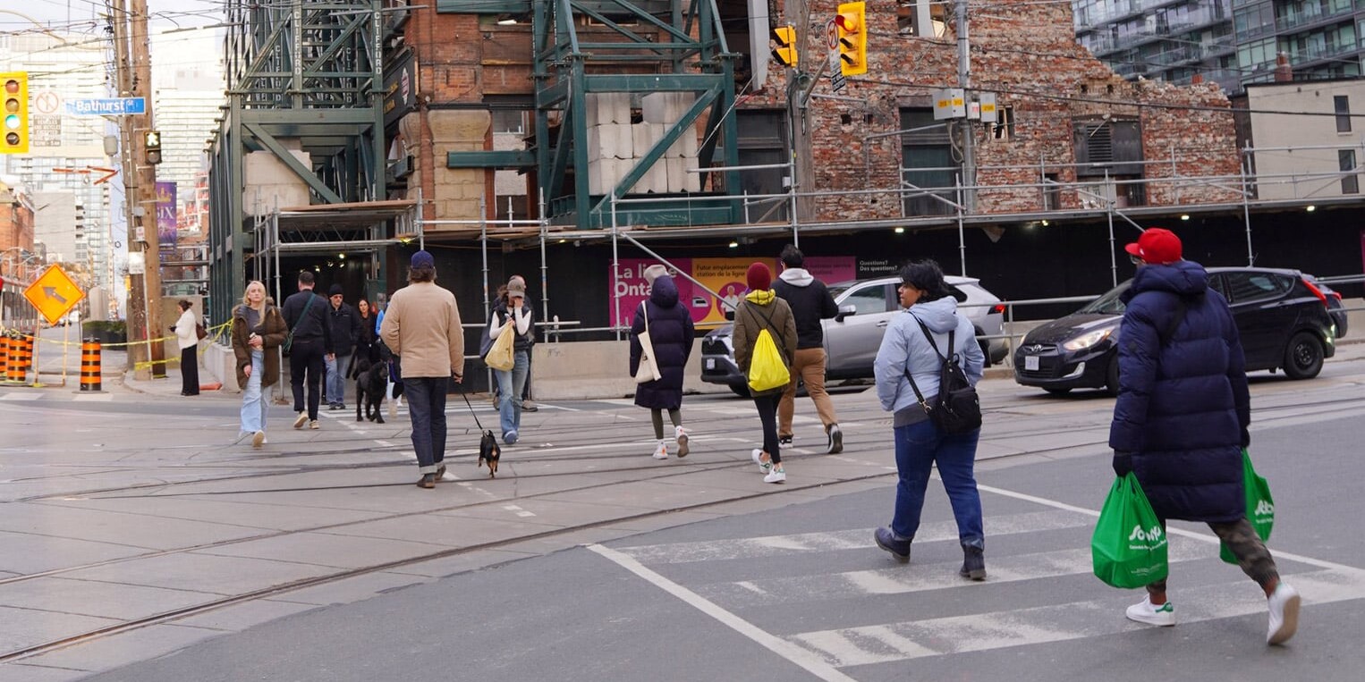 A group of people cross a city street at a crosswalk near a construction site, some carrying green shopping bags. Buildings and traffic lights are visible, reflecting how Canadians aren’t just adapting to diversity – there are data to show we’re embracing it.
