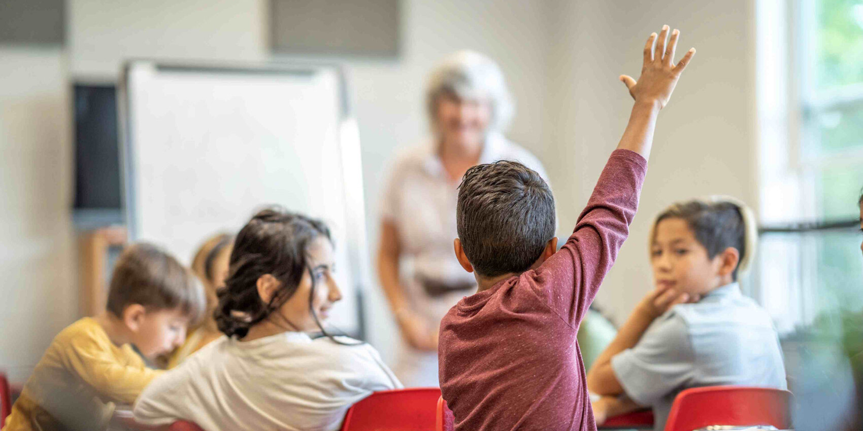 A young student, part of refugees to Canada, raises his hand in a classroom, while other children sit at red chairs and a teacher stands near a whiteboard in the background—a snapshot of schools and teachers shaping new beginnings.