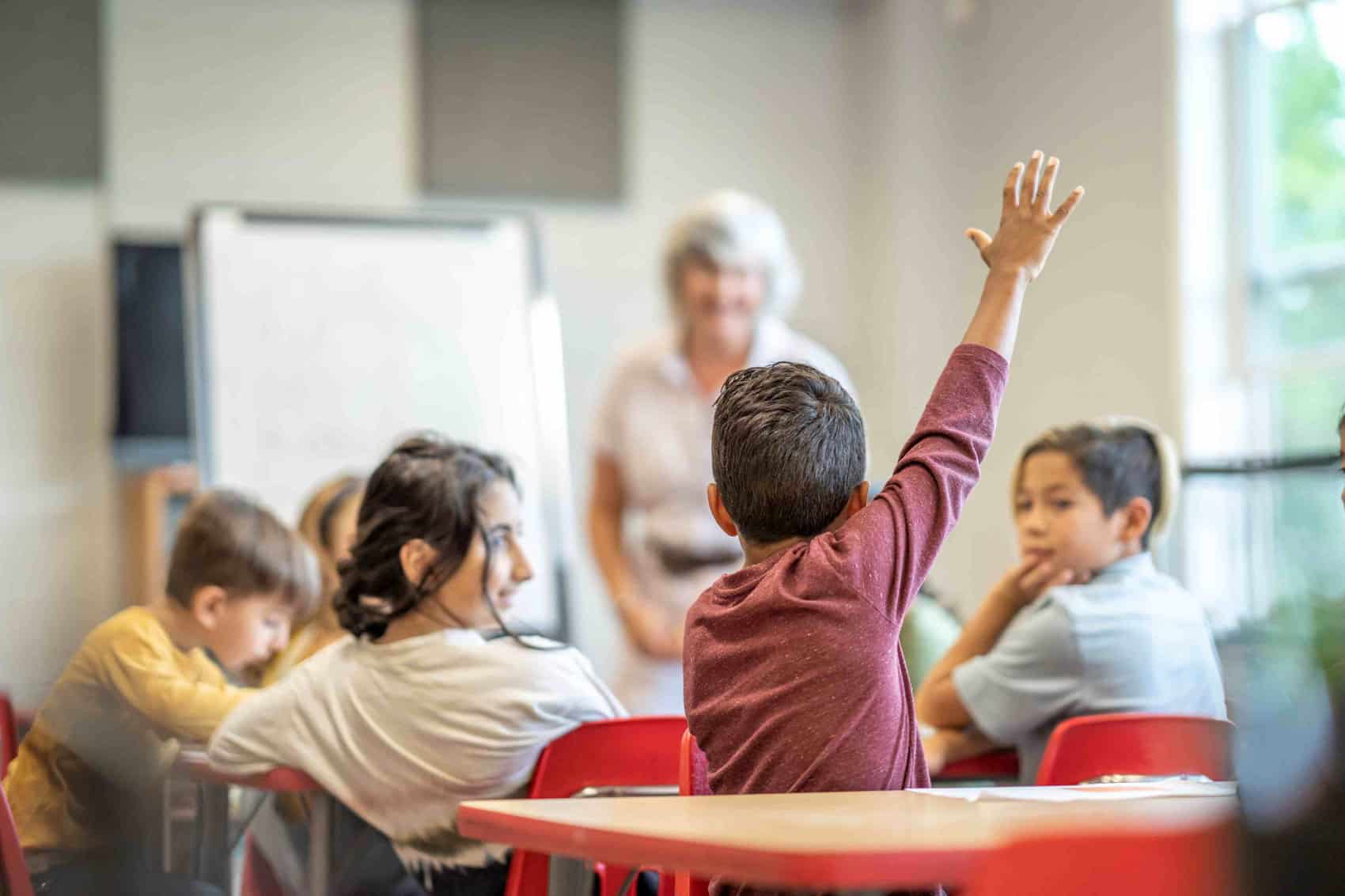 A young student, part of refugees to Canada, raises his hand in a classroom, while other children sit at red chairs and a teacher stands near a whiteboard in the background—a snapshot of schools and teachers shaping new beginnings.