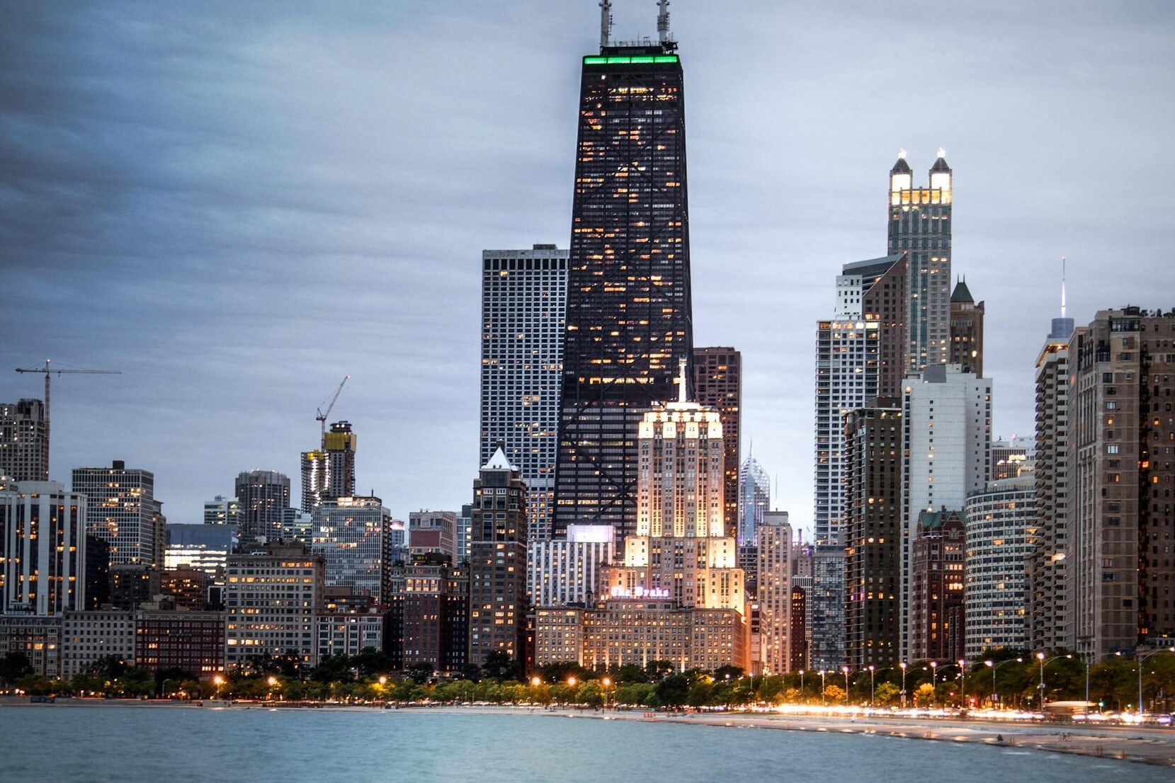 A city skyline at dusk with tall buildings illuminated, including a black skyscraper with green lights on top, bordered by smaller buildings, trees, and water—reflecting one issue on which Canadians aren't polarized: their views of the U.S.