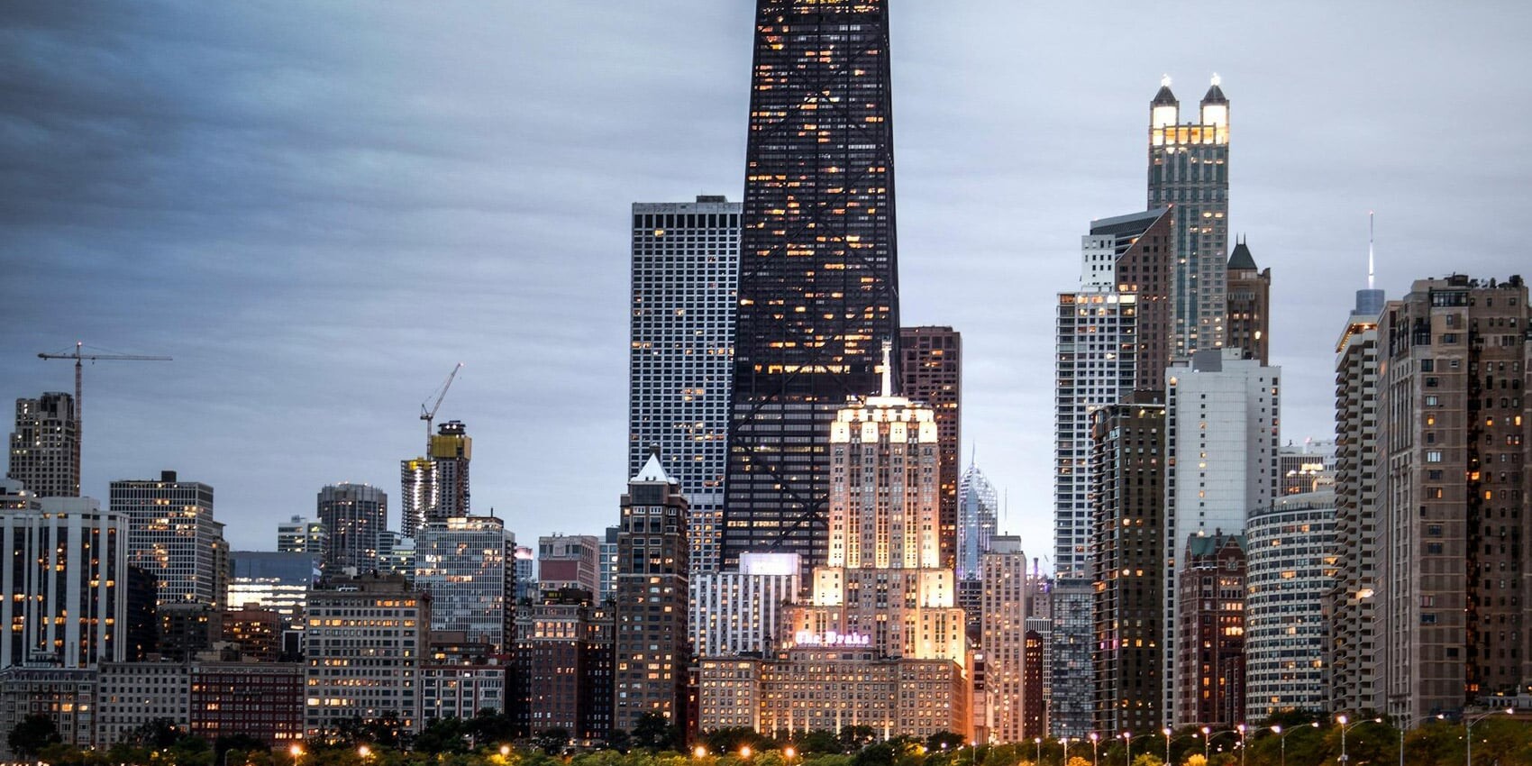 A city skyline at dusk with tall buildings illuminated, including a black skyscraper with green lights on top, bordered by smaller buildings, trees, and water—reflecting one issue on which Canadians aren't polarized: their views of the U.S.