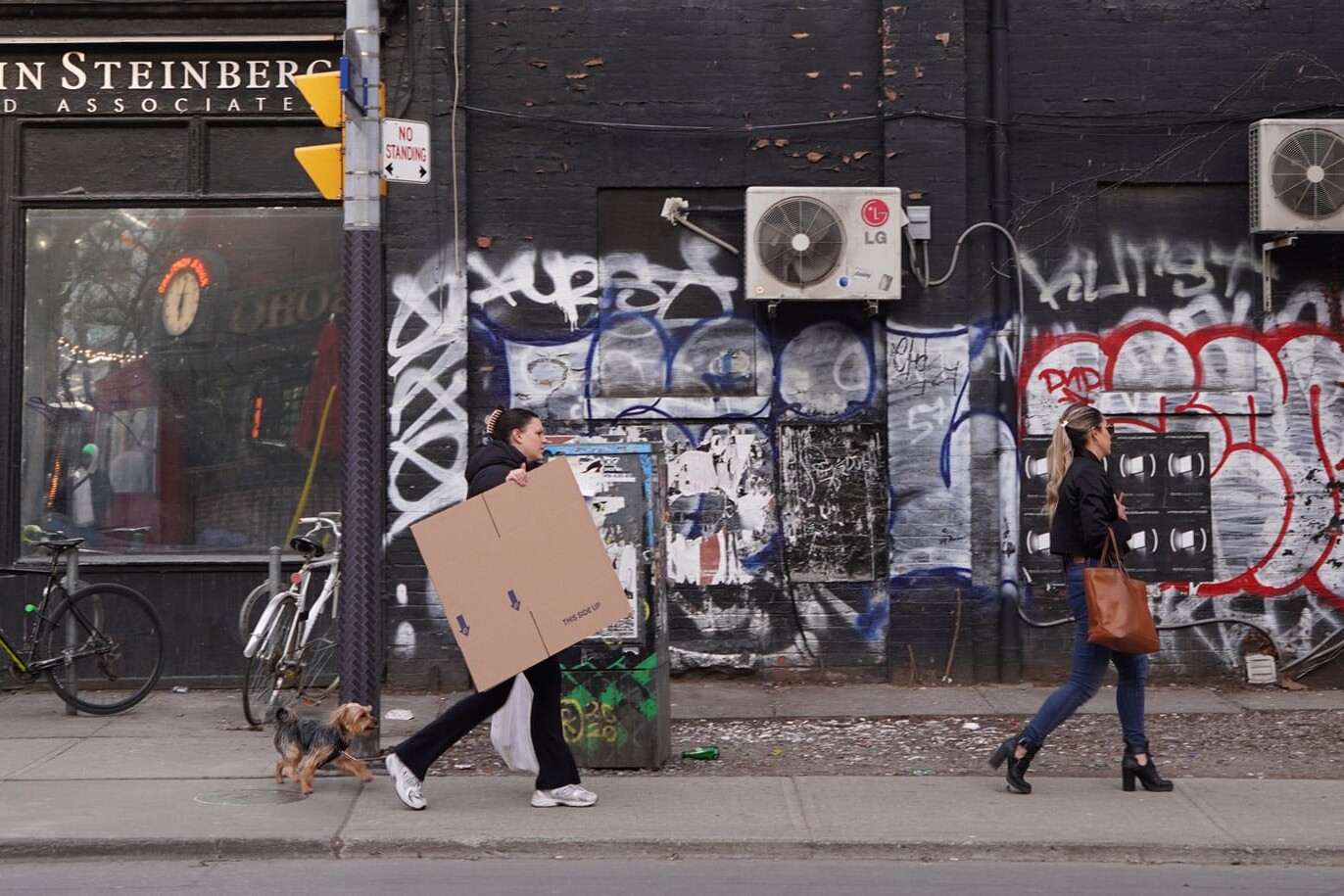 Two women walk along a graffiti-covered street; one carries a large cardboard box with a small dog on a leash, while the other, ahead, wears heels and a black coat. Bicycles, air conditioners, and “Canada is sorry – a lot” are scrawled on the wall.