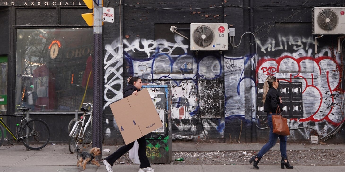 Two women walk along a graffiti-covered street; one carries a large cardboard box with a small dog on a leash, while the other, ahead, wears heels and a black coat. Bicycles, air conditioners, and “Canada is sorry – a lot” are scrawled on the wall.