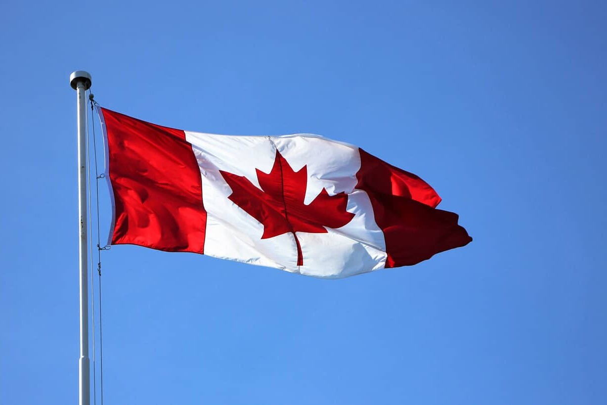The Canadian flag, featuring a red maple leaf on white with red bars, waves proudly on a flagpole against a clear blue sky—a fitting symbol for the nation known for its politeness, as Canada is sorry – a lot.