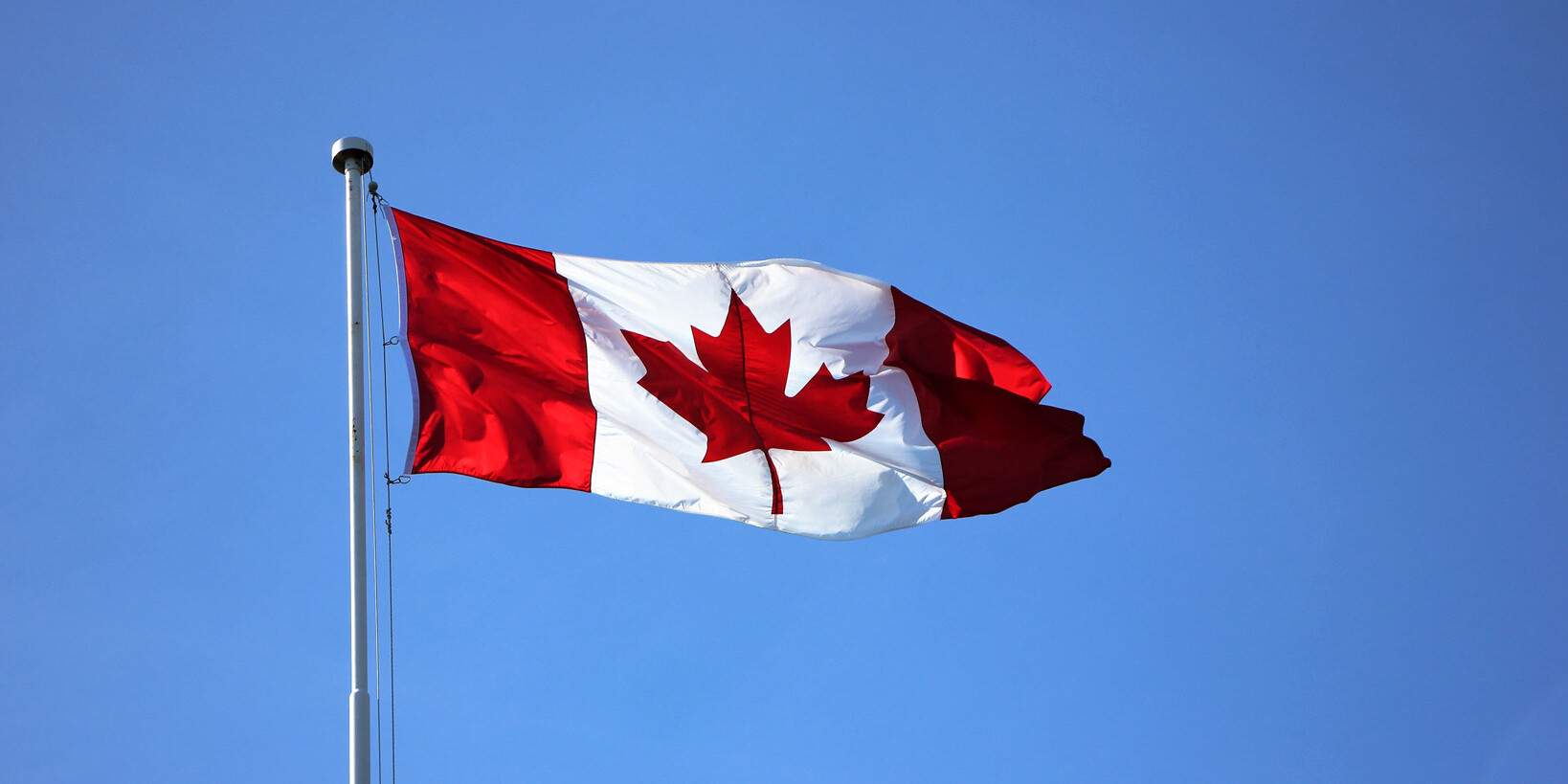 The Canadian flag, featuring a red maple leaf on white with red bars, waves proudly on a flagpole against a clear blue sky—a fitting symbol for the nation known for its politeness, as Canada is sorry – a lot.