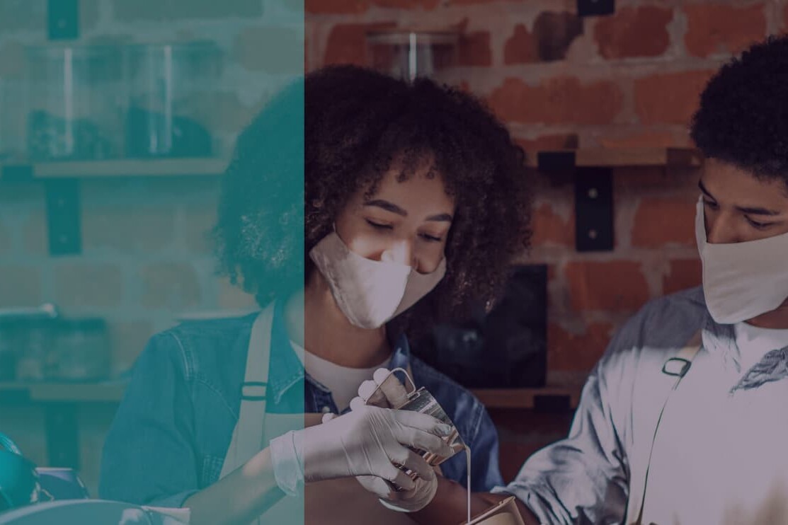 Two people wearing face masks and gloves collaborate with glass containers in a cozy, brick-walled workspace, highlighting participation in skills training. Shelves and jars line the background, suggesting a hands-on learning environment.
