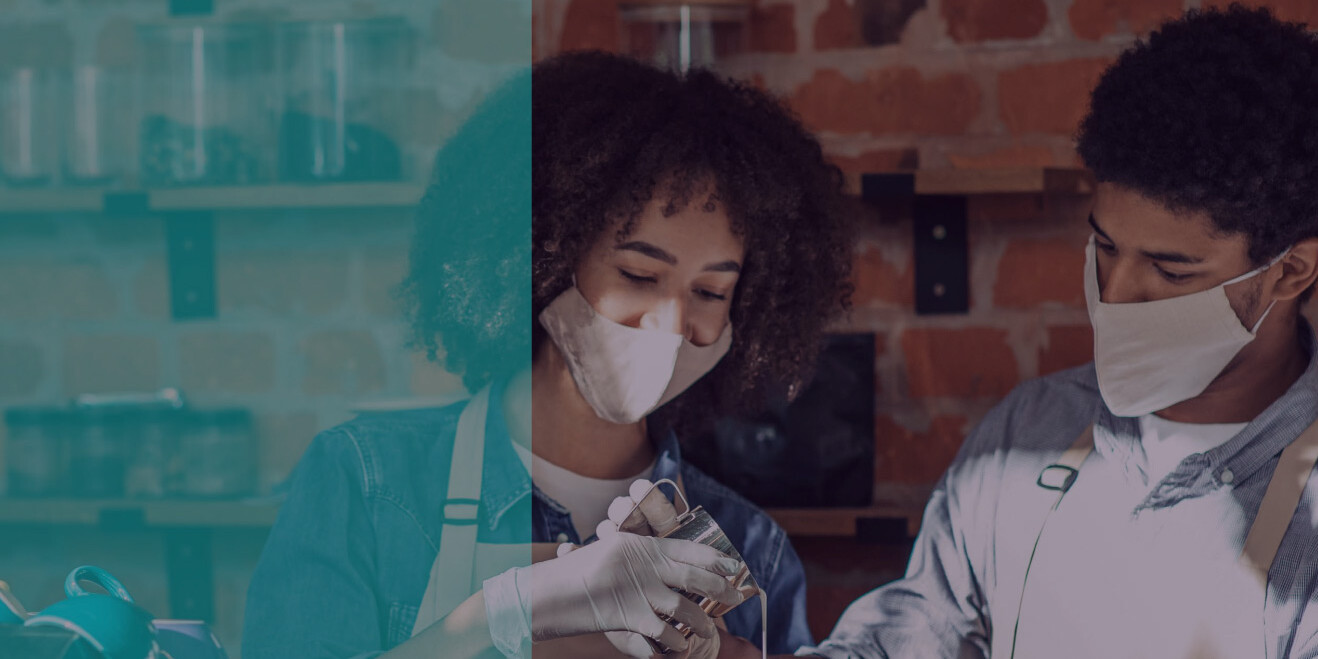 Two people wearing face masks and gloves collaborate with glass containers in a cozy, brick-walled workspace, highlighting participation in skills training. Shelves and jars line the background, suggesting a hands-on learning environment.