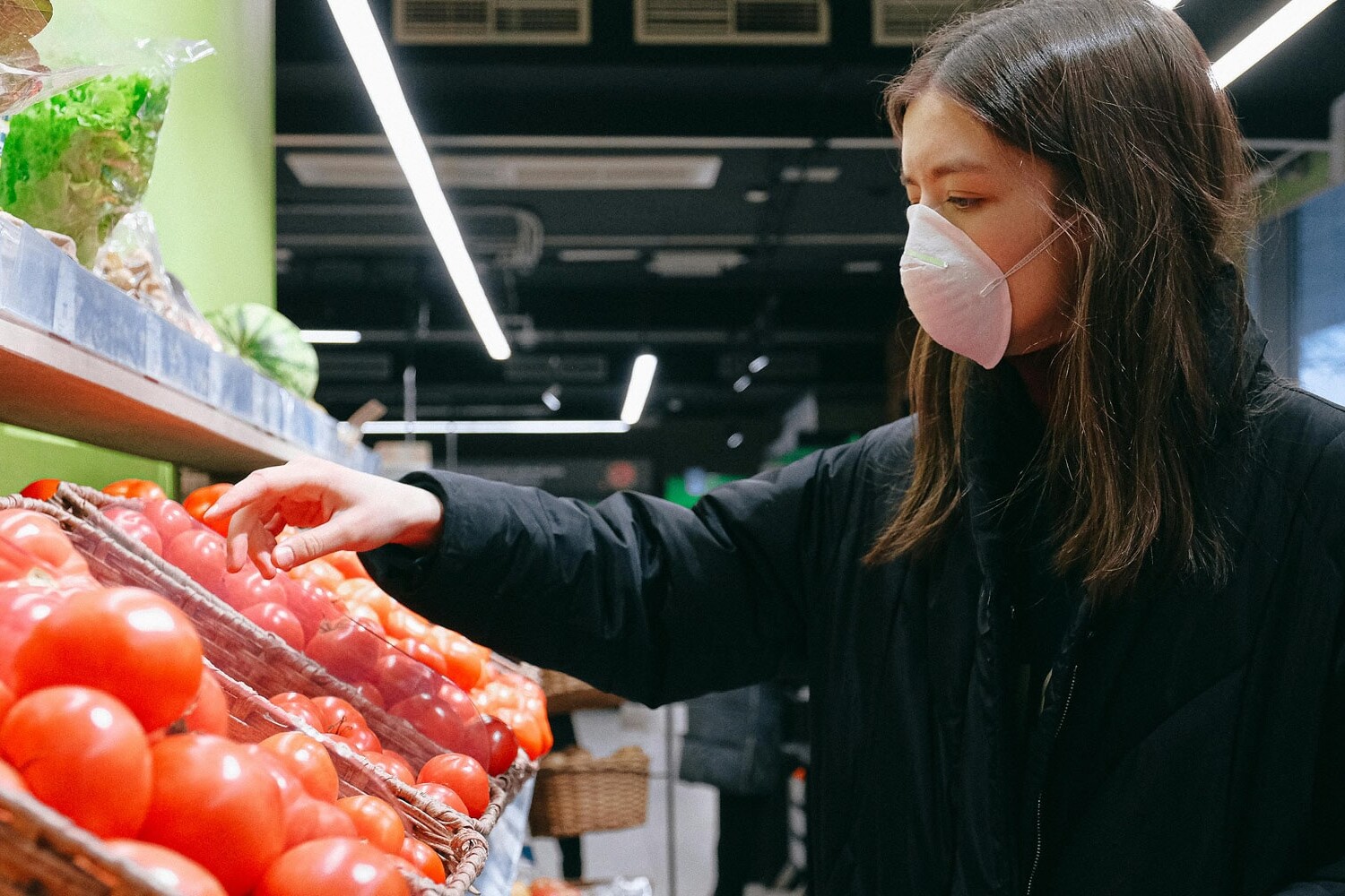 A woman wearing a mask and dark coat selects tomatoes from a display at a grocery store, reflecting how Canadians are sharply more dissatisfied with direction of country at end of 2023, as she stands by shelves of fresh vegetables under bright indoor lighting.