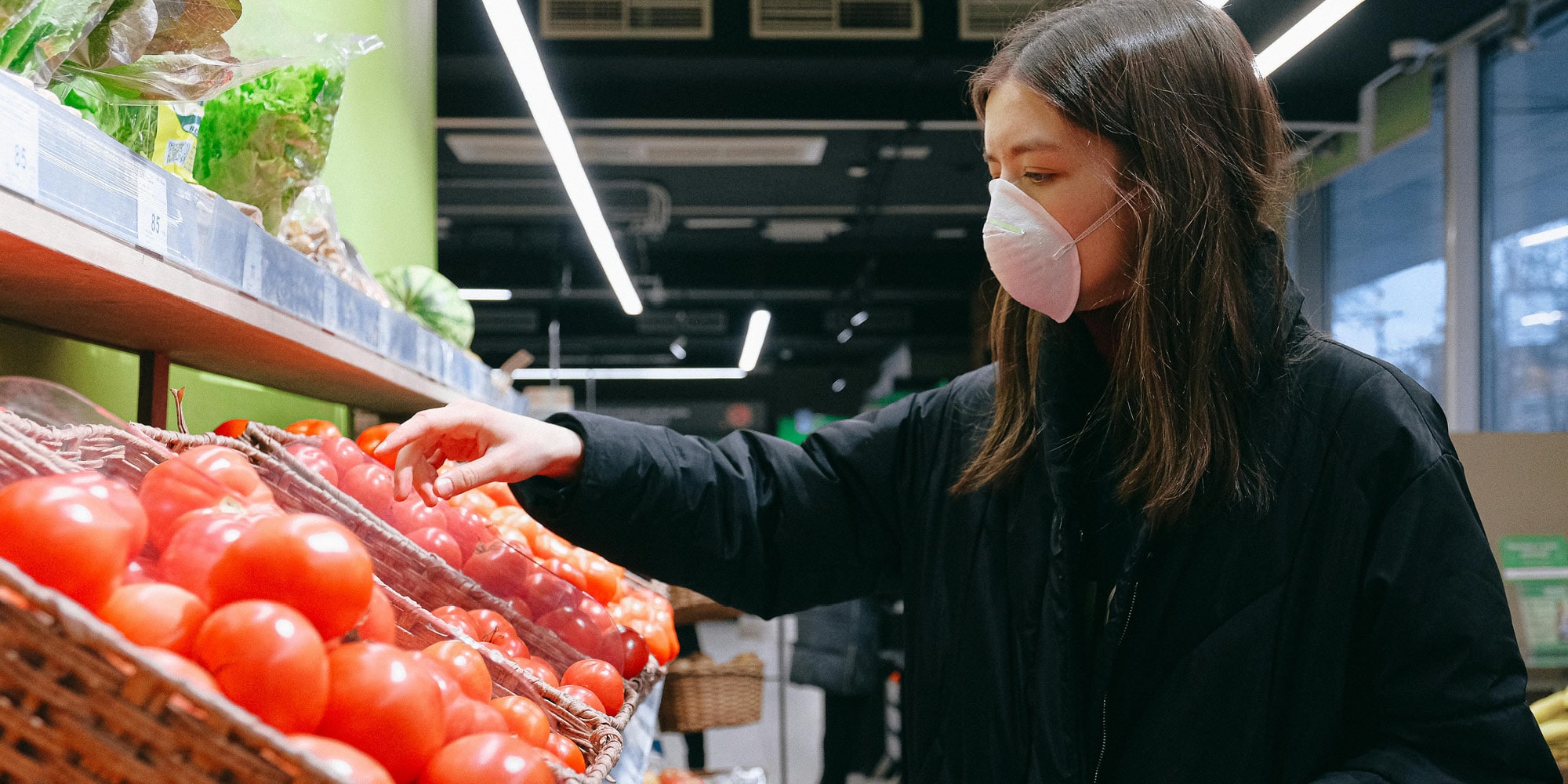 A woman wearing a mask and dark coat selects tomatoes from a display at a grocery store, reflecting how Canadians are sharply more dissatisfied with direction of country at end of 2023, as she stands by shelves of fresh vegetables under bright indoor lighting.