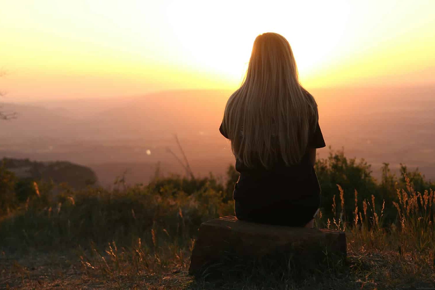 A person with long hair sits on a rock, facing a bright sunset over a scenic landscape with hills and grass, embodying the peaceful atmosphere that surveys show Canadians experience more than their often more polarized and angry American neighbors.