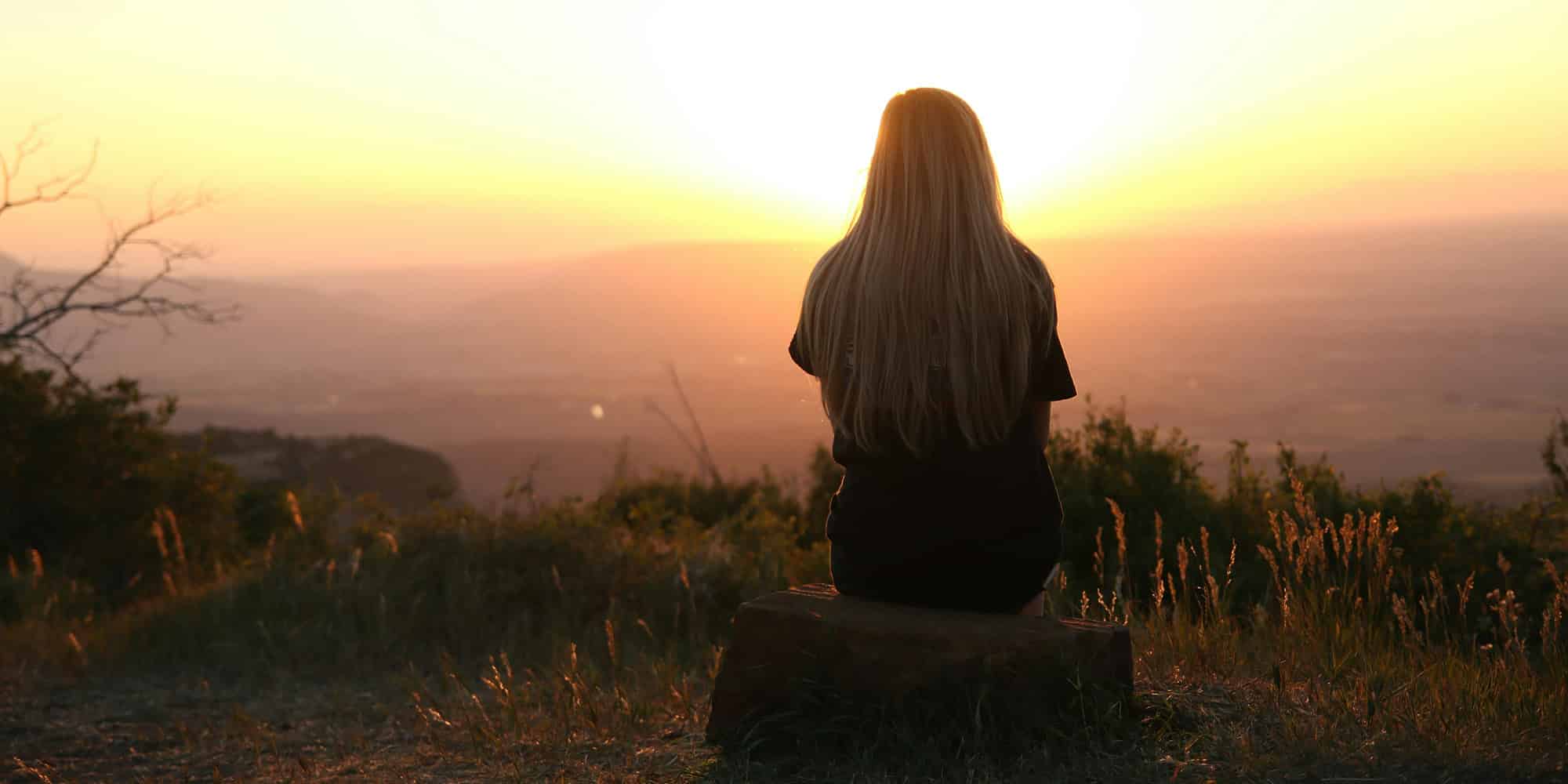 A person with long hair sits on a rock, facing a bright sunset over a scenic landscape with hills and grass, embodying the peaceful atmosphere that surveys show Canadians experience more than their often more polarized and angry American neighbors.
