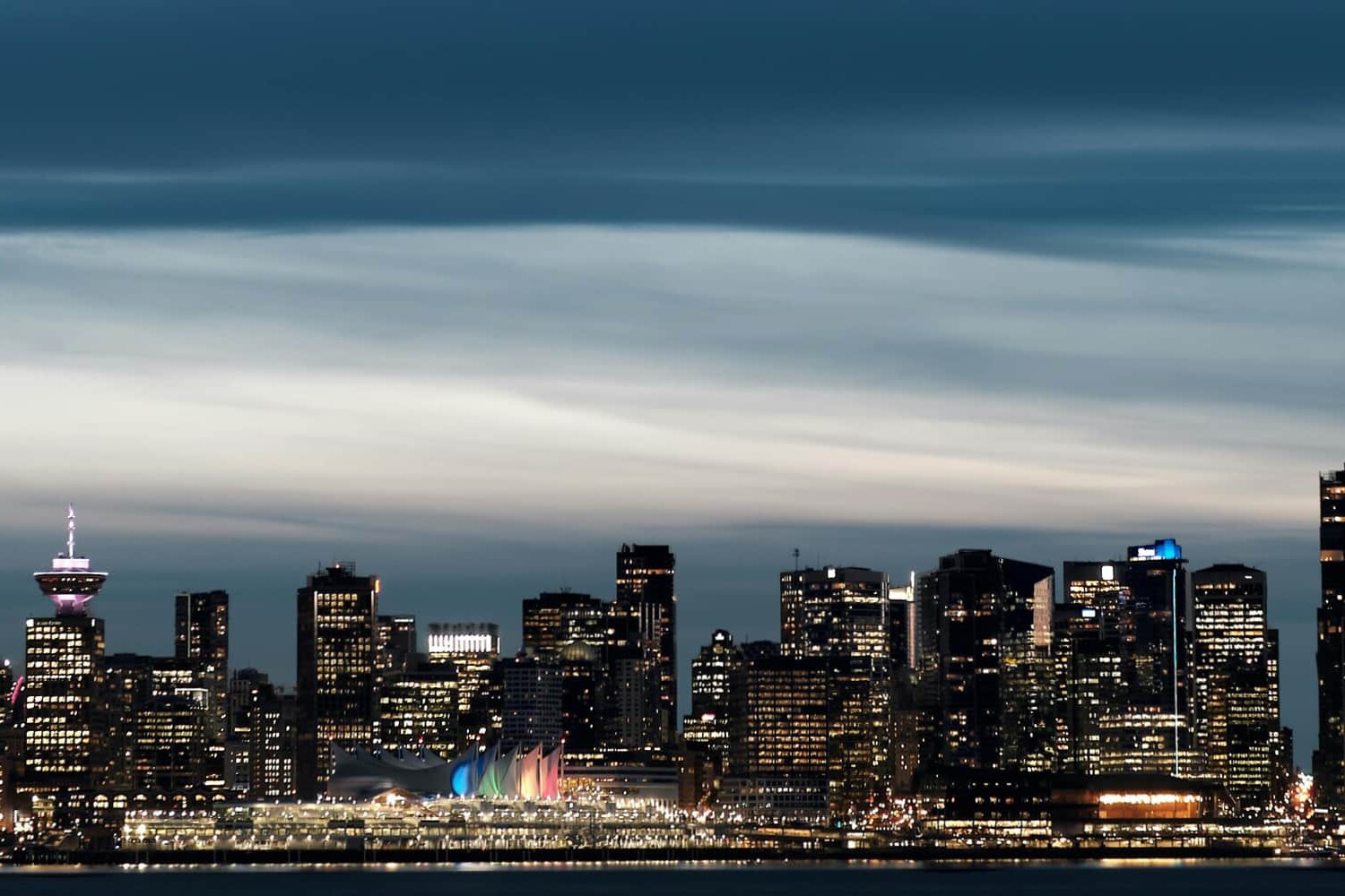 A nighttime cityscape of Vancouver, Canada, featuring illuminated skyscrapers, the Canada Place building with its colorful sails, and a dramatic, cloudy sky—a stunning backdrop often seen in discussions about Canadian politics.