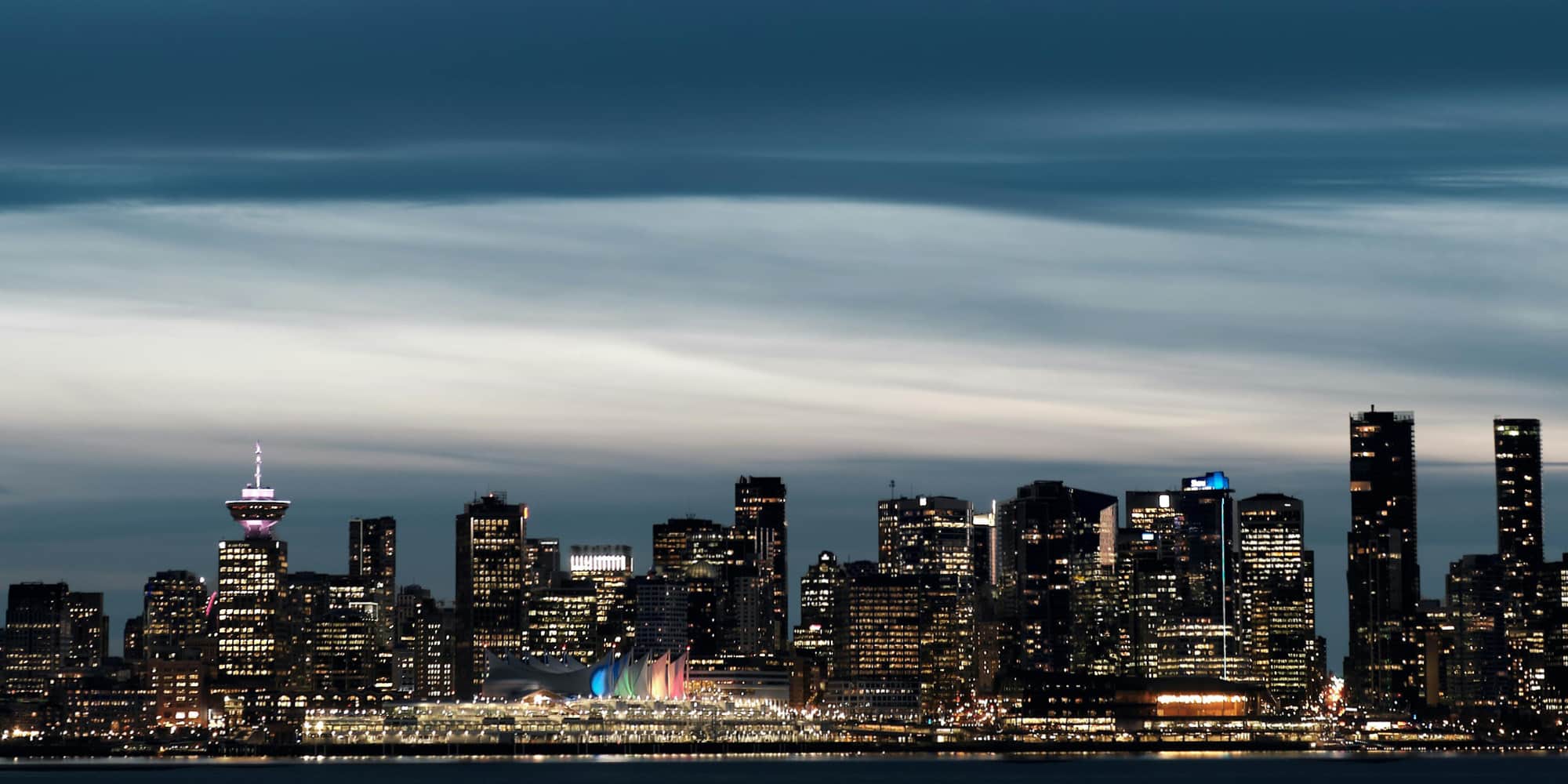 A nighttime cityscape of Vancouver, Canada, featuring illuminated skyscrapers, the Canada Place building with its colorful sails, and a dramatic, cloudy sky—a stunning backdrop often seen in discussions about Canadian politics.