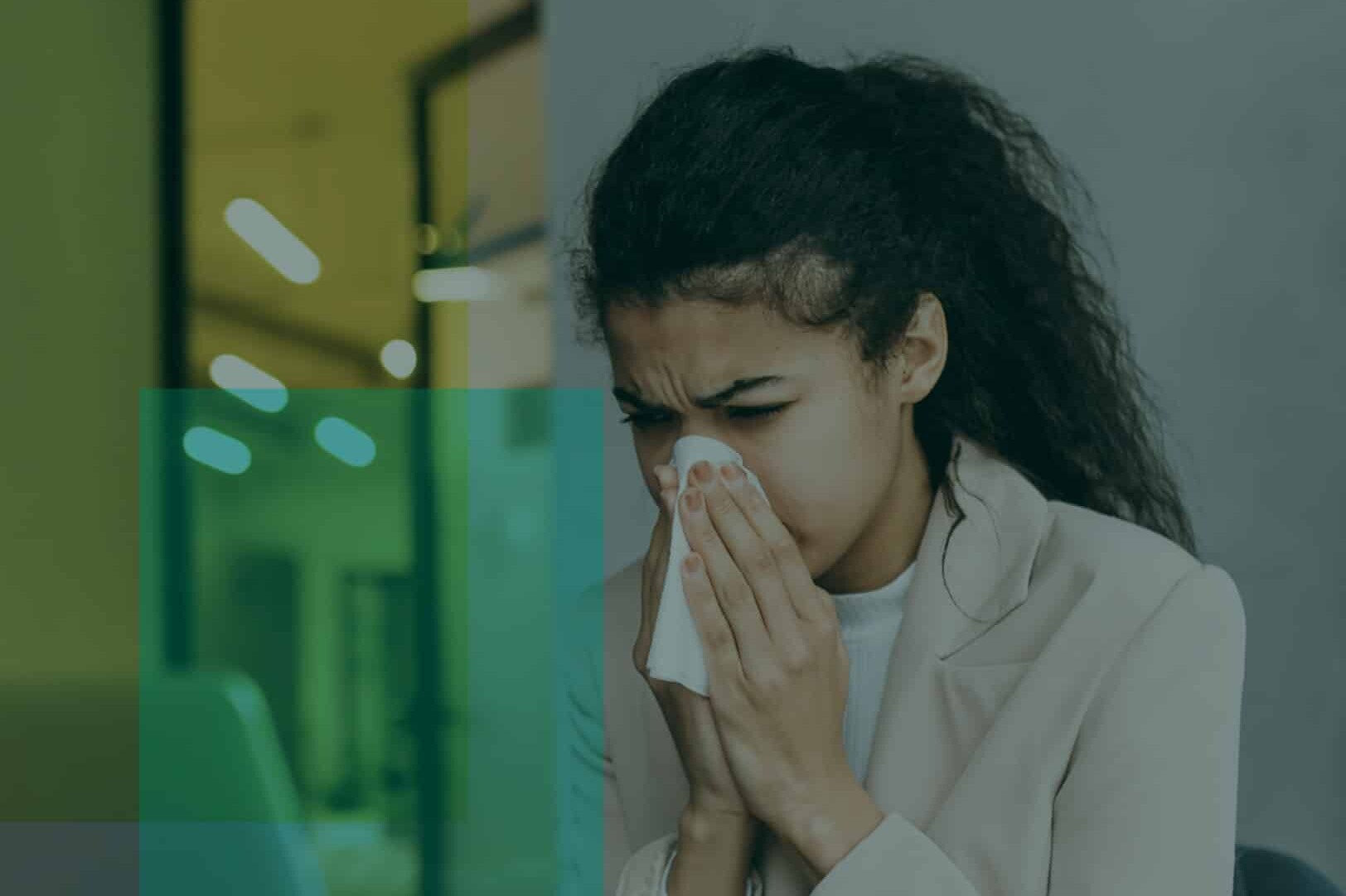 A woman with long dark hair is sitting indoors, holding a tissue to her nose, appearing to sneeze or have a cold. She is wearing a light-colored blazer and looks uncomfortable, possibly experiencing the lingering effects of COVID-19.