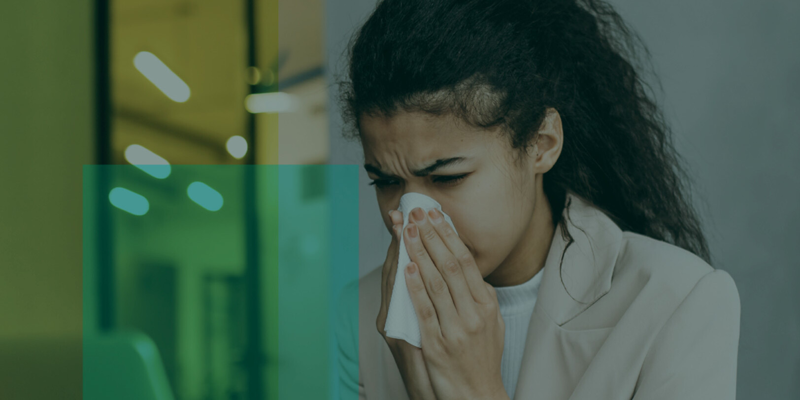 A woman with long dark hair is sitting indoors, holding a tissue to her nose, appearing to sneeze or have a cold. She is wearing a light-colored blazer and looks uncomfortable, possibly experiencing the lingering effects of COVID-19.