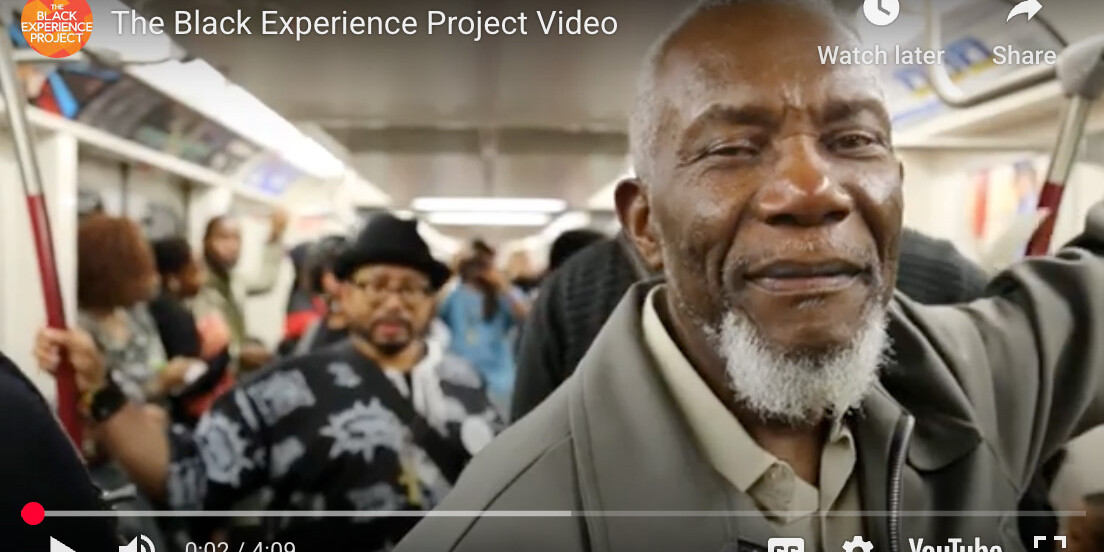A smiling older man with a gray beard stands in the foreground of a crowded subway train. Other passengers, including a man in a patterned shirt and hat, are visible behind him. The Black Experience Project captures this vibrant moment.