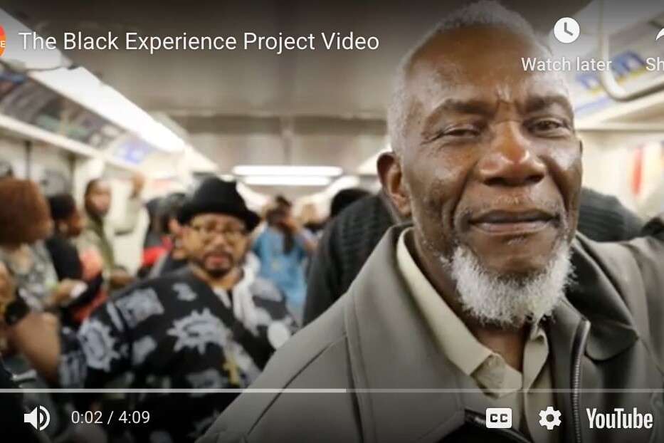 A smiling older man with a gray beard stands in the foreground of a crowded subway train. Other passengers, including a man in a patterned shirt and hat, are visible behind him. The Black Experience Project captures this vibrant moment.