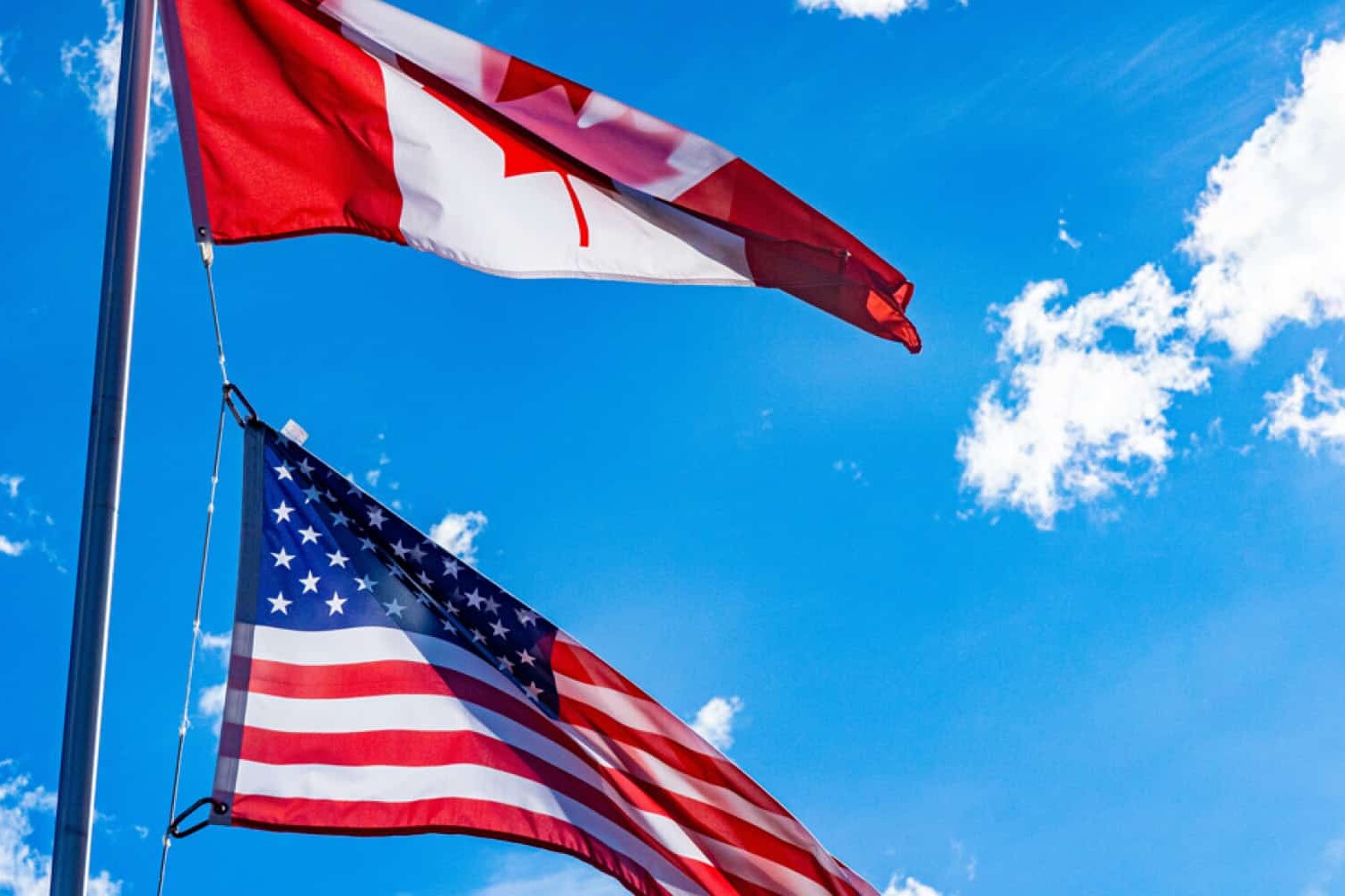 Two flags, the Canadian flag above and the American flag below, flutter on a flagpole against a bright blue sky—a striking symbol of Canada and the United States' political and cultural connections.
