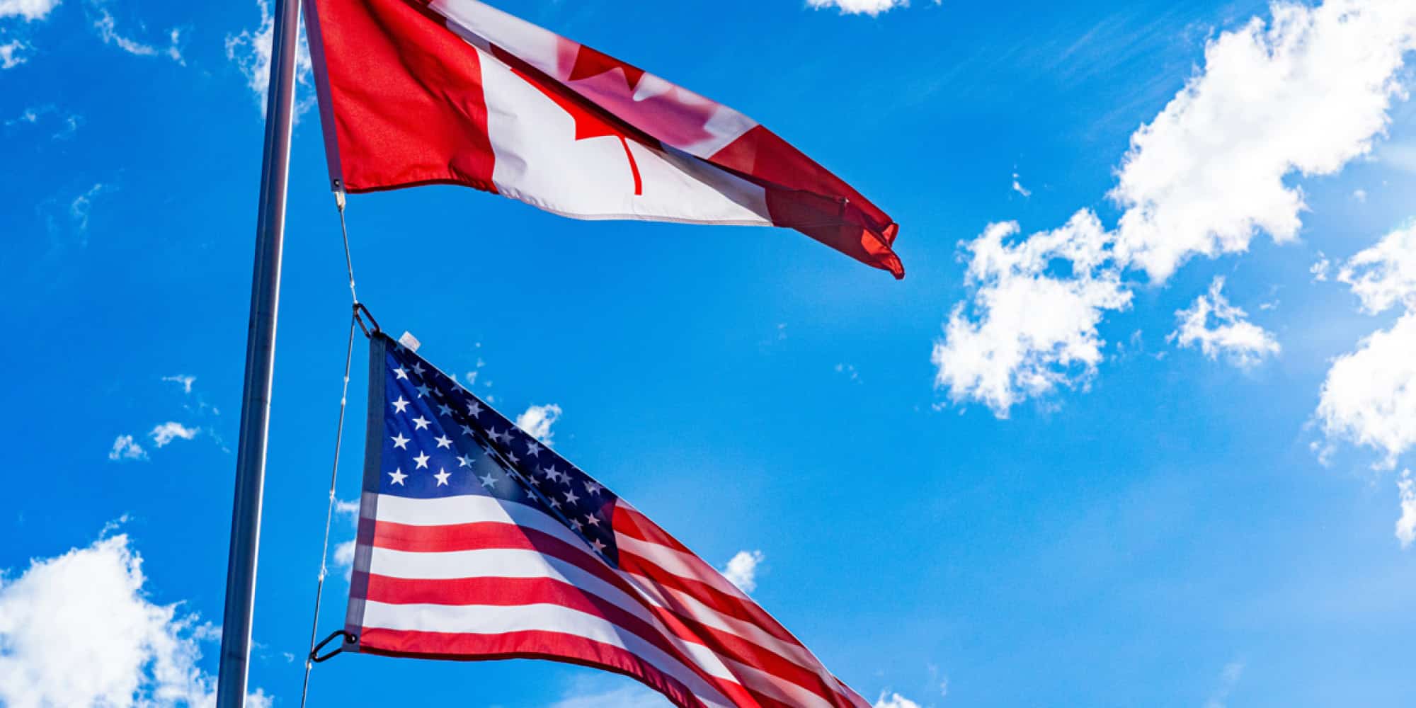 Two flags, the Canadian flag above and the American flag below, flutter on a flagpole against a bright blue sky—a striking symbol of Canada and the United States' political and cultural connections.