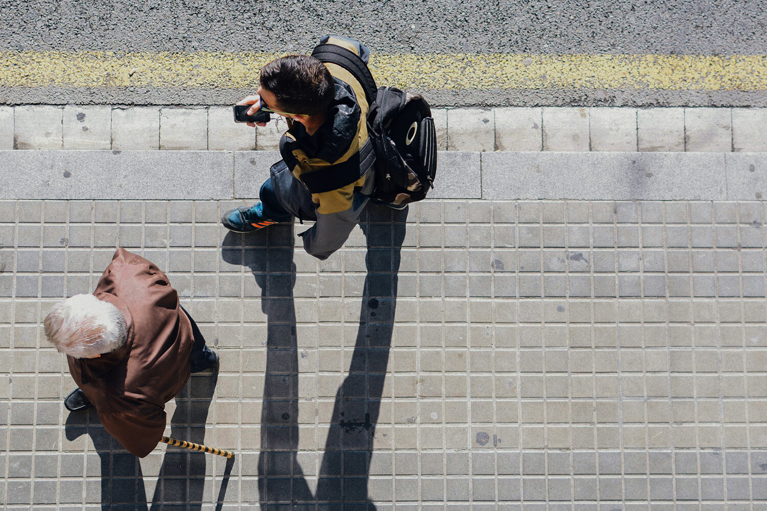 Overhead view of an elderly person with a cane walking on a sidewalk alongside a younger person carrying a backpack, both casting long shadows—reflecting the generational perspectives in five public-opinion trends to watch in 2025.