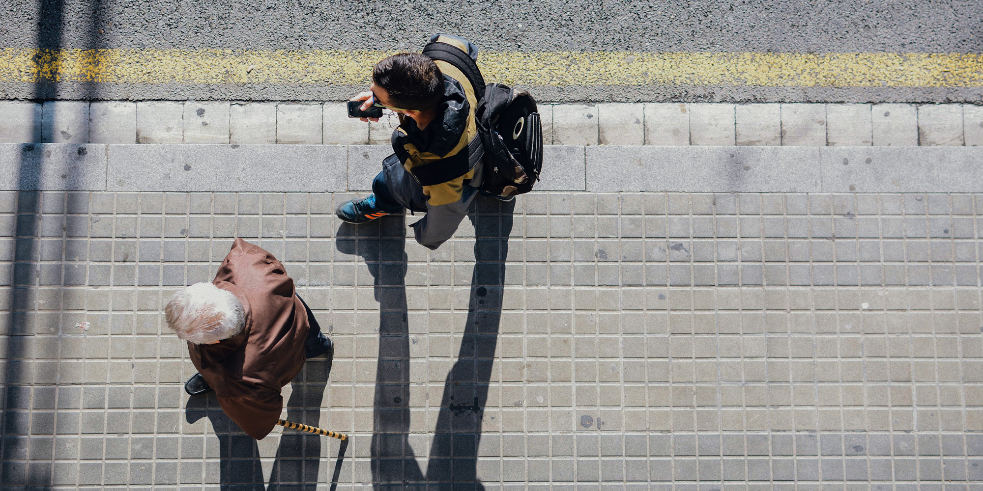 Overhead view of an elderly person with a cane walking on a sidewalk alongside a younger person carrying a backpack, both casting long shadows—reflecting the generational perspectives in five public-opinion trends to watch in 2025.