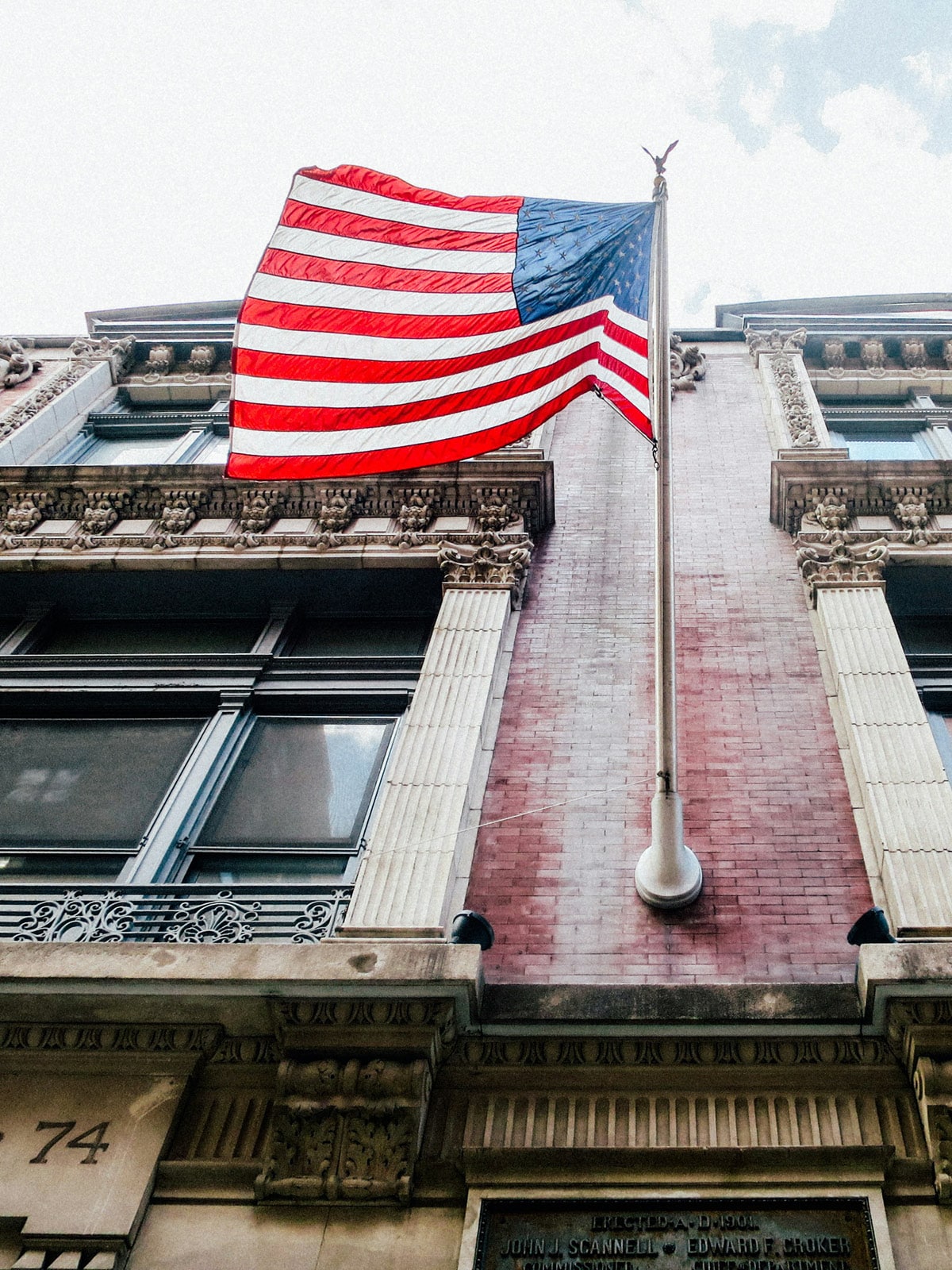 The American flag waves atop a tall flagpole mounted on the facade of a historic brick and stone building, seen from a low-angle perspective against a partly cloudy sky, symbolizing the political and cultural ties between Canada and the United States.