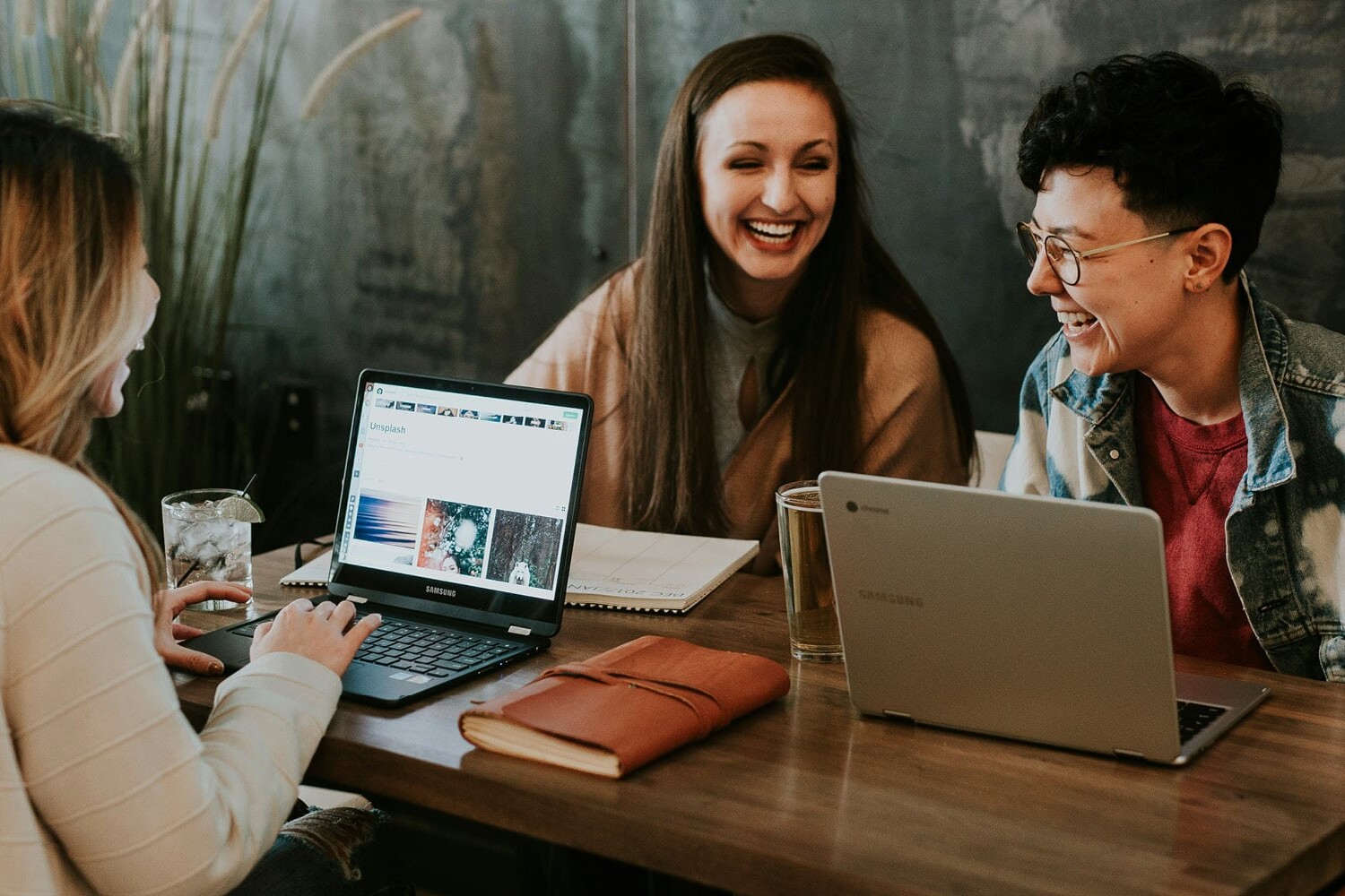 Three people sit at a wooden table with laptops, notebooks, and drinks, smiling and laughing together in a casual, cozy setting that reflects diversity, equity and inclusion.