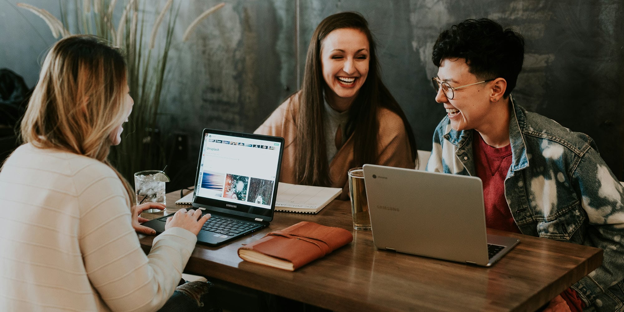 Three people sit at a wooden table with laptops, notebooks, and drinks, smiling and laughing together in a casual, cozy setting that reflects diversity, equity and inclusion.