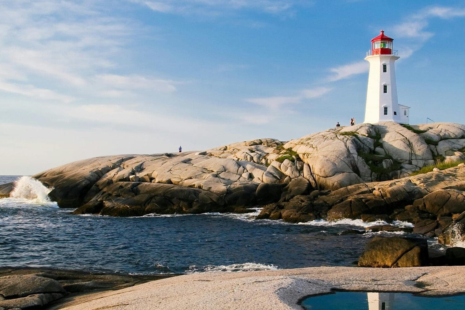 A white lighthouse with a red top stands on smooth gray rocks by the ocean under a blue sky. Waves crash nearby, and a few people linger—our elbows may be up but have Canadians really changed in these timeless coastal moments?.