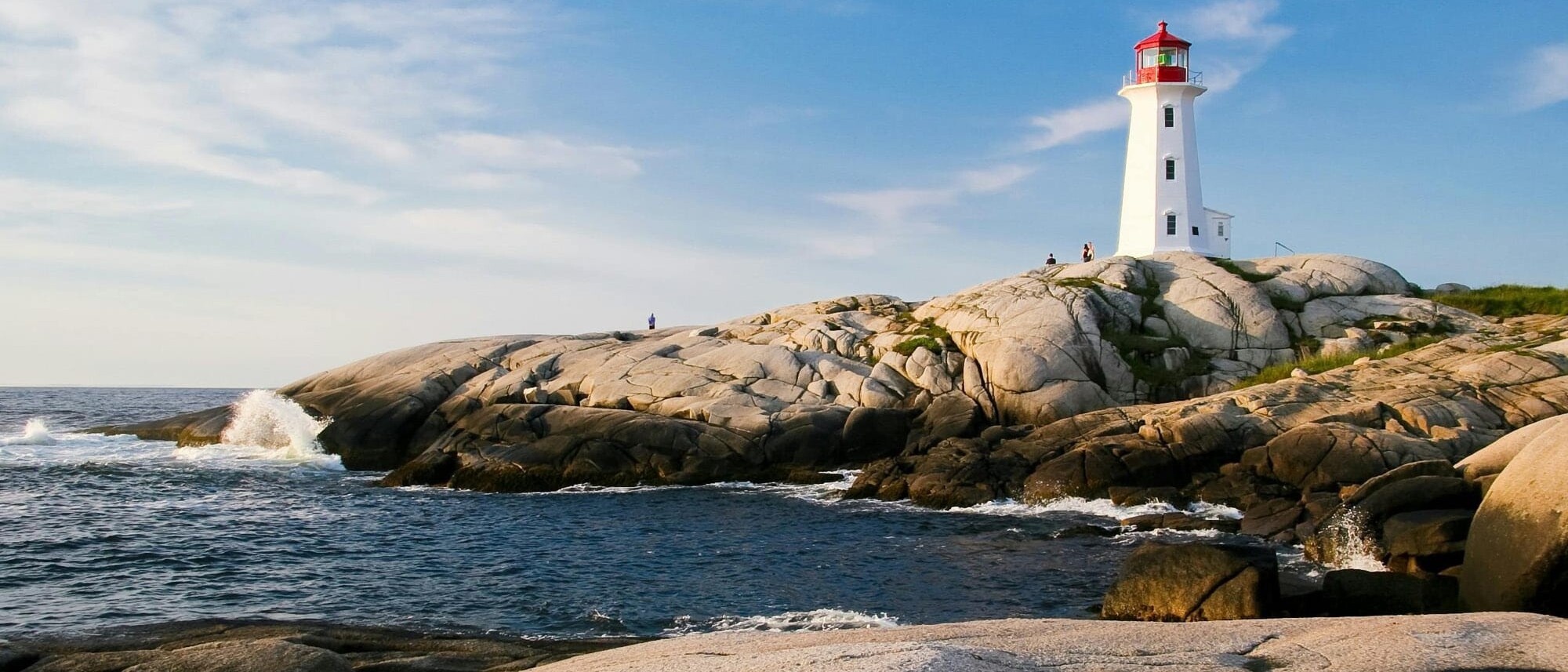 A white lighthouse with a red top stands on smooth gray rocks by the ocean under a blue sky. Waves crash nearby, and a few people linger—our elbows may be up but have Canadians really changed in these timeless coastal moments?.