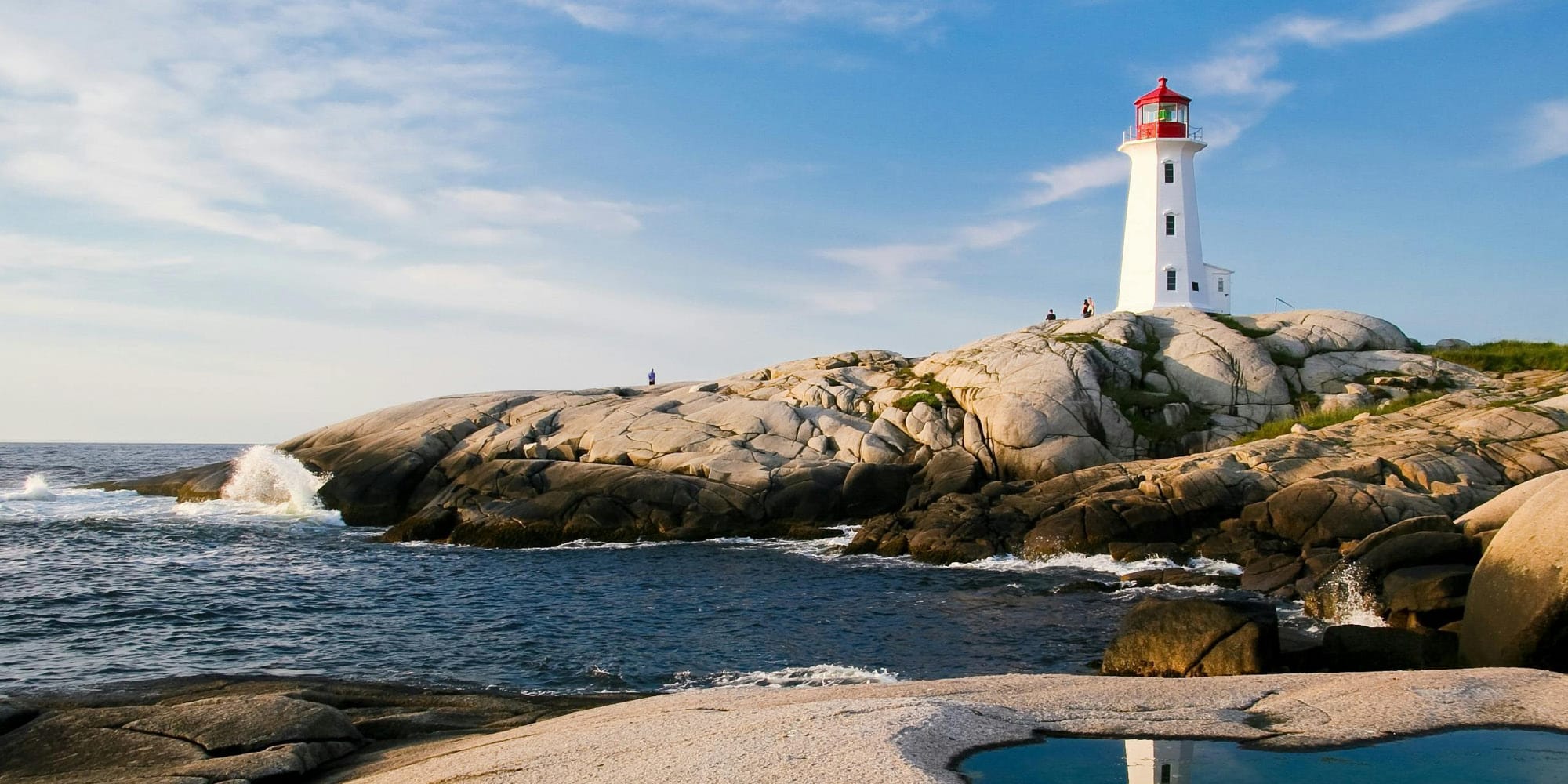 A white lighthouse with a red top stands on smooth gray rocks by the ocean under a blue sky. Waves crash nearby, and a few people linger—our elbows may be up but have Canadians really changed in these timeless coastal moments?.