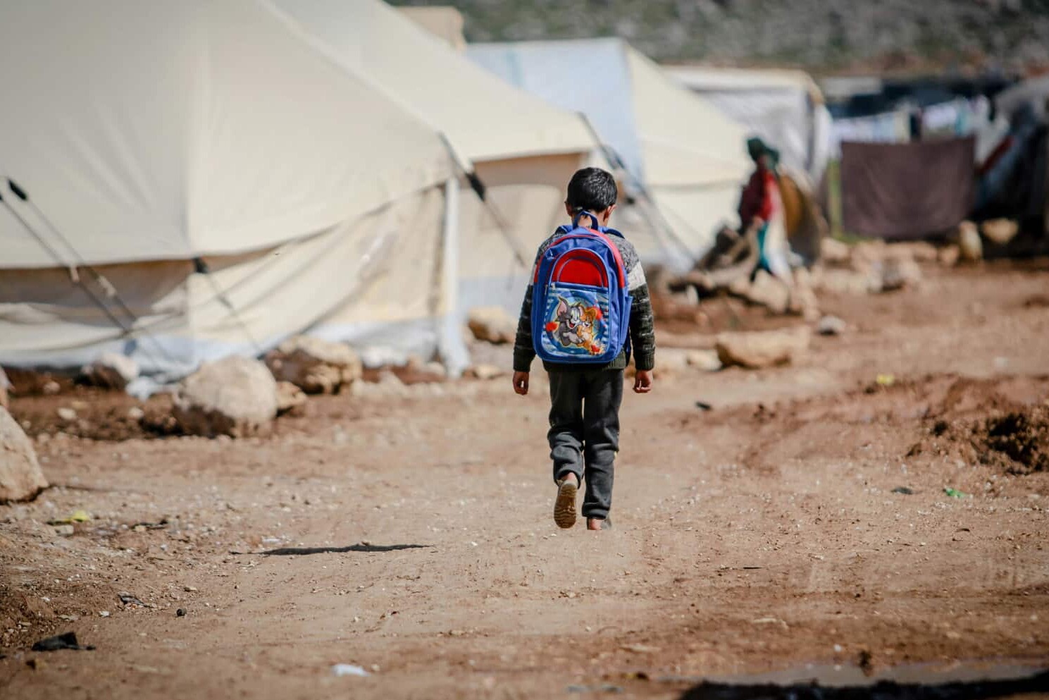 A child with a blue backpack walks alone on a dirt path between tents in a refugee camp, surrounded by rocks and makeshift shelters—each step writing a new immigration story for Canada.