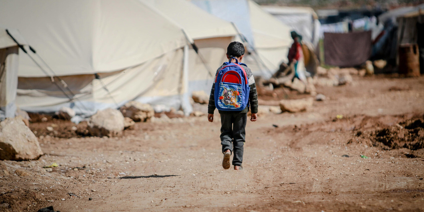 A child with a blue backpack walks alone on a dirt path between tents in a refugee camp, surrounded by rocks and makeshift shelters—each step writing a new immigration story for Canada.
