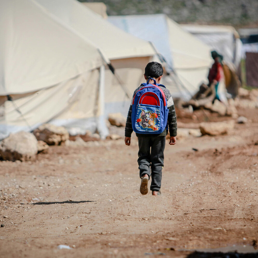 A child with a blue backpack walks alone on a dirt path between tents in a refugee camp, surrounded by rocks and makeshift shelters—each step writing a new immigration story for Canada.